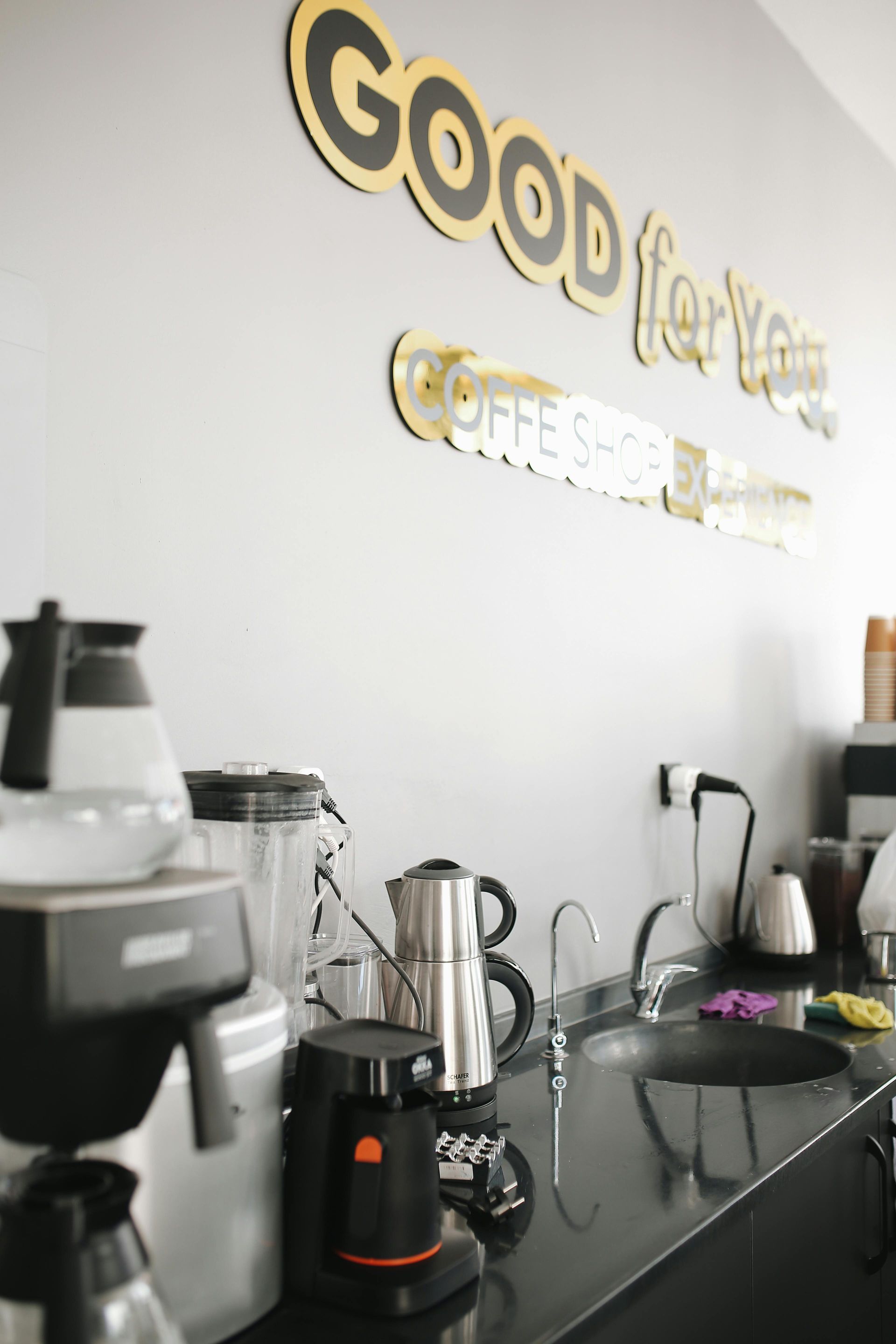Coffee station with countertop machines and kettles beneath a “GOOD” wall sign