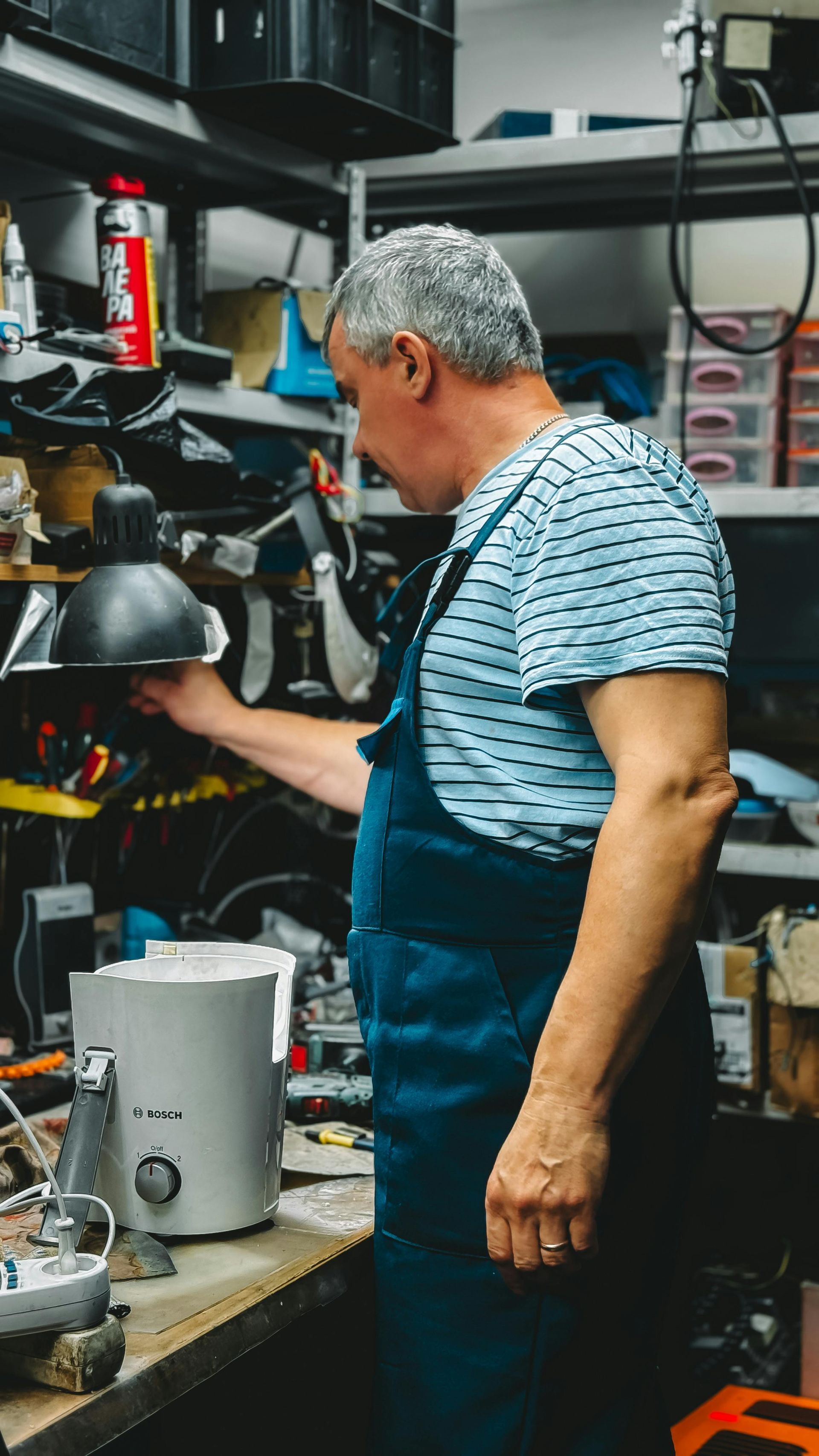 Person in a workshop using a tool at a cluttered workbench, wearing a striped shirt and apron.