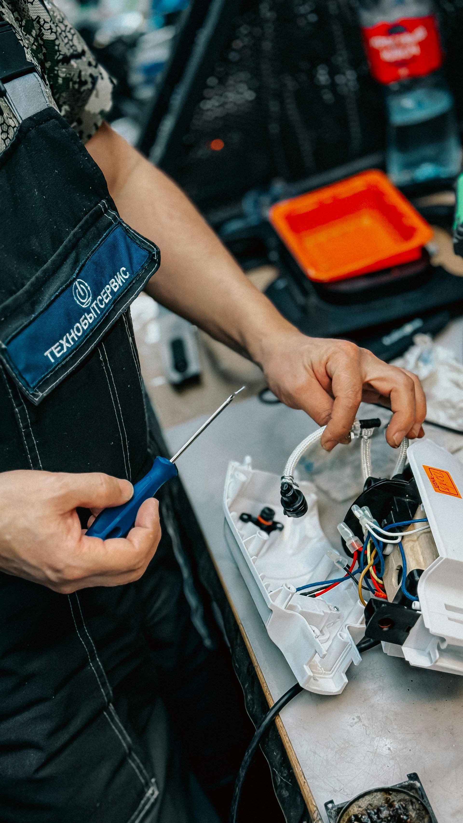 Person holding a blue screwdriver near a disassembled device on a workbench with tools and wires