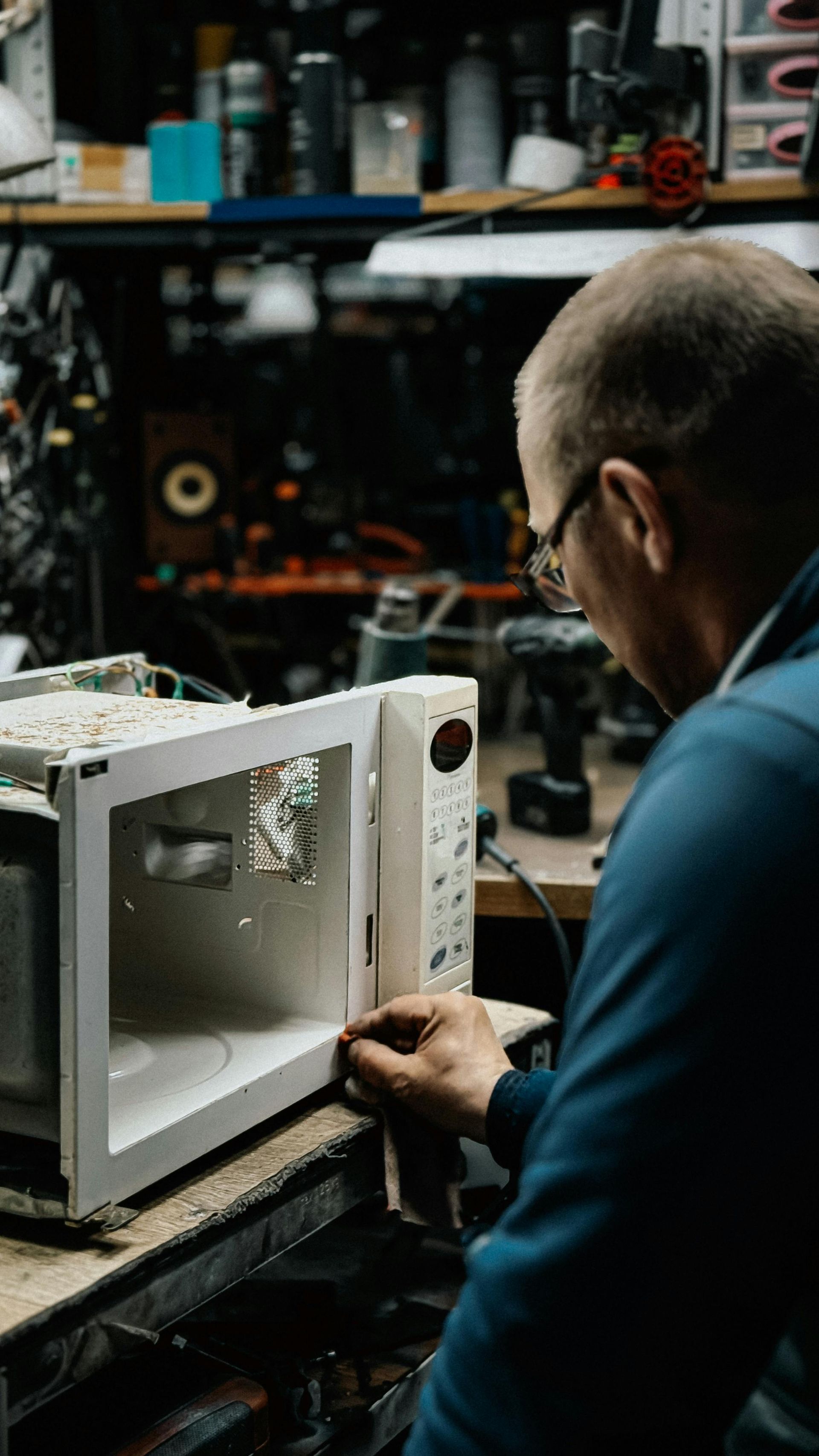Person inspecting a white microwave in a cluttered workshop with tools and equipment in the background