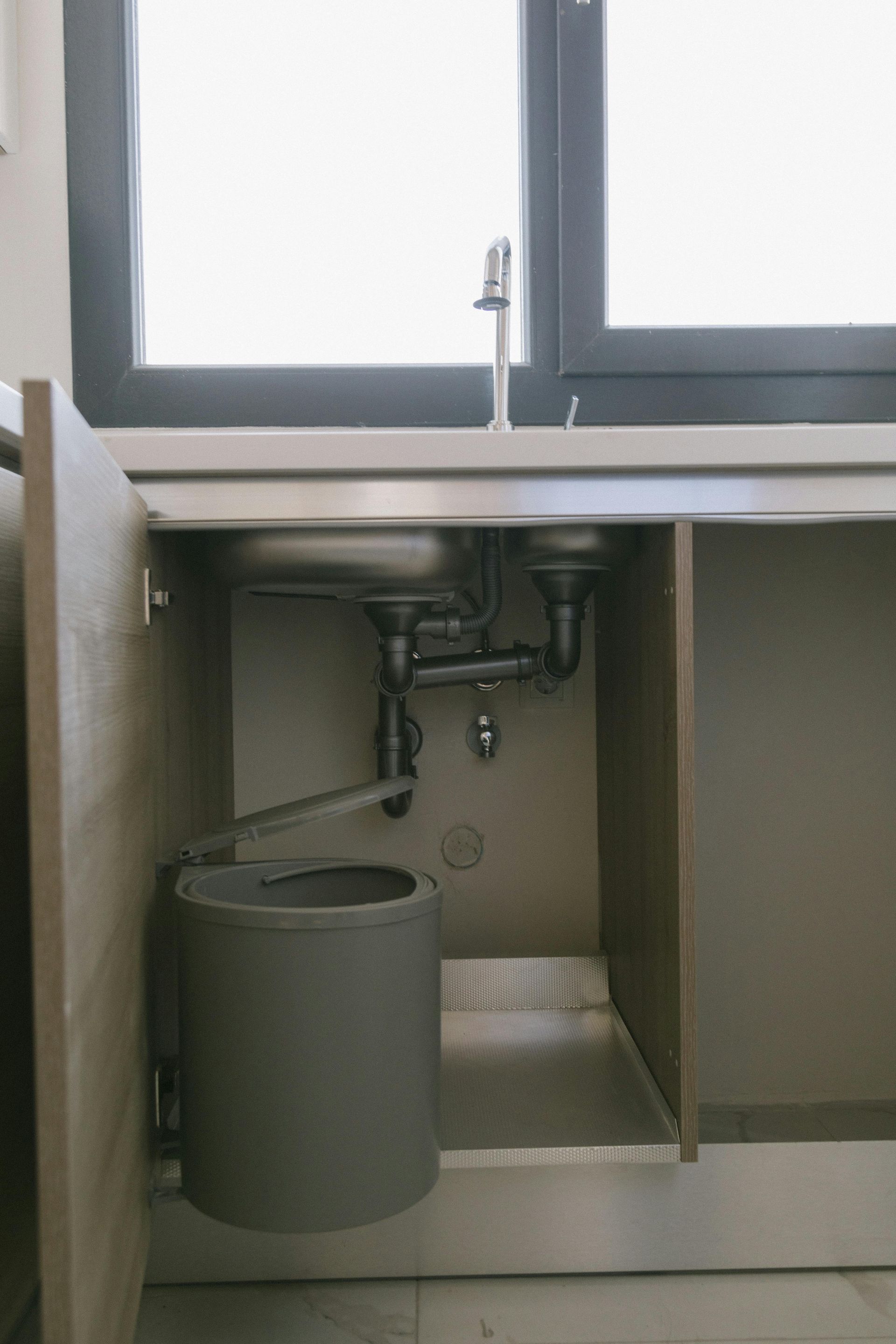 Small bathroom sink area with a window above, exposed plumbing, and a gray trash bin underneath.