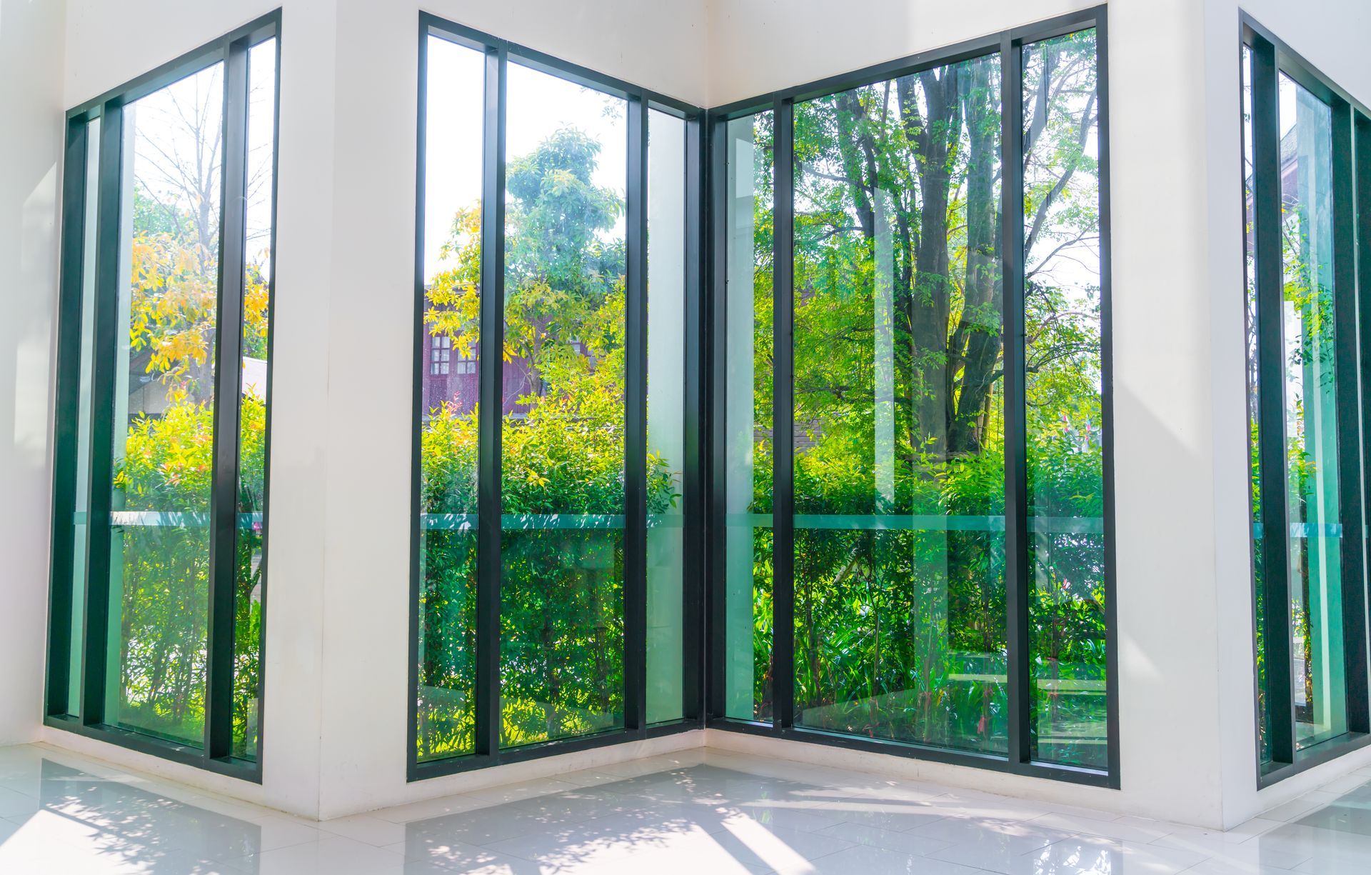 Bright corner windows with black frames overlook greenery and yellow flowers in a sunlit room