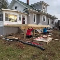 Two men are working on a wooden deck in front of a house.