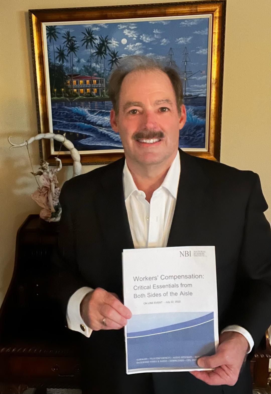 A man in a suit is holding a book in front of a painting.