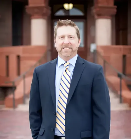 Man in navy suit and striped tie, smiles in front of a red brick building with columns.
