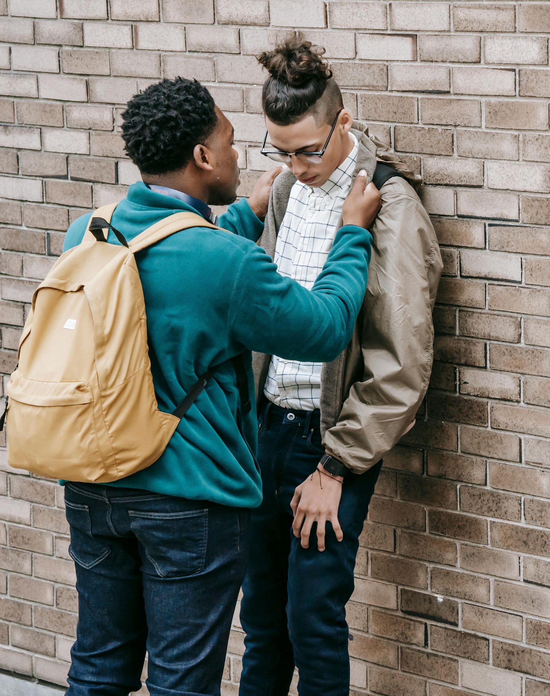 Two young men, one adjusting the other’s jacket near a brick wall. One wears a teal sweatshirt, the other a tan jacket.
