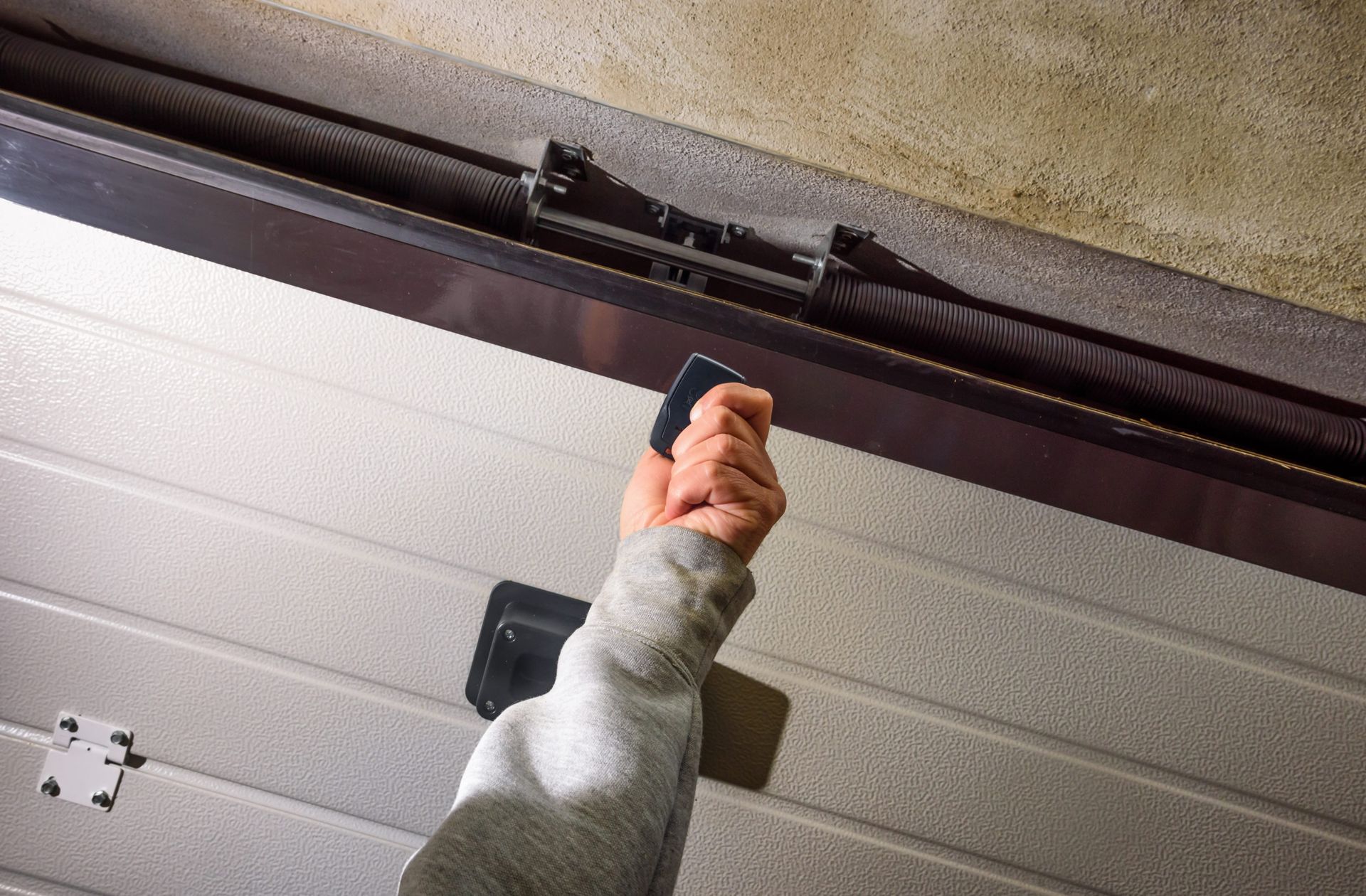 Person's hand using a garage door opener remote. The garage door is partially open, and the opener mechanism is visible.