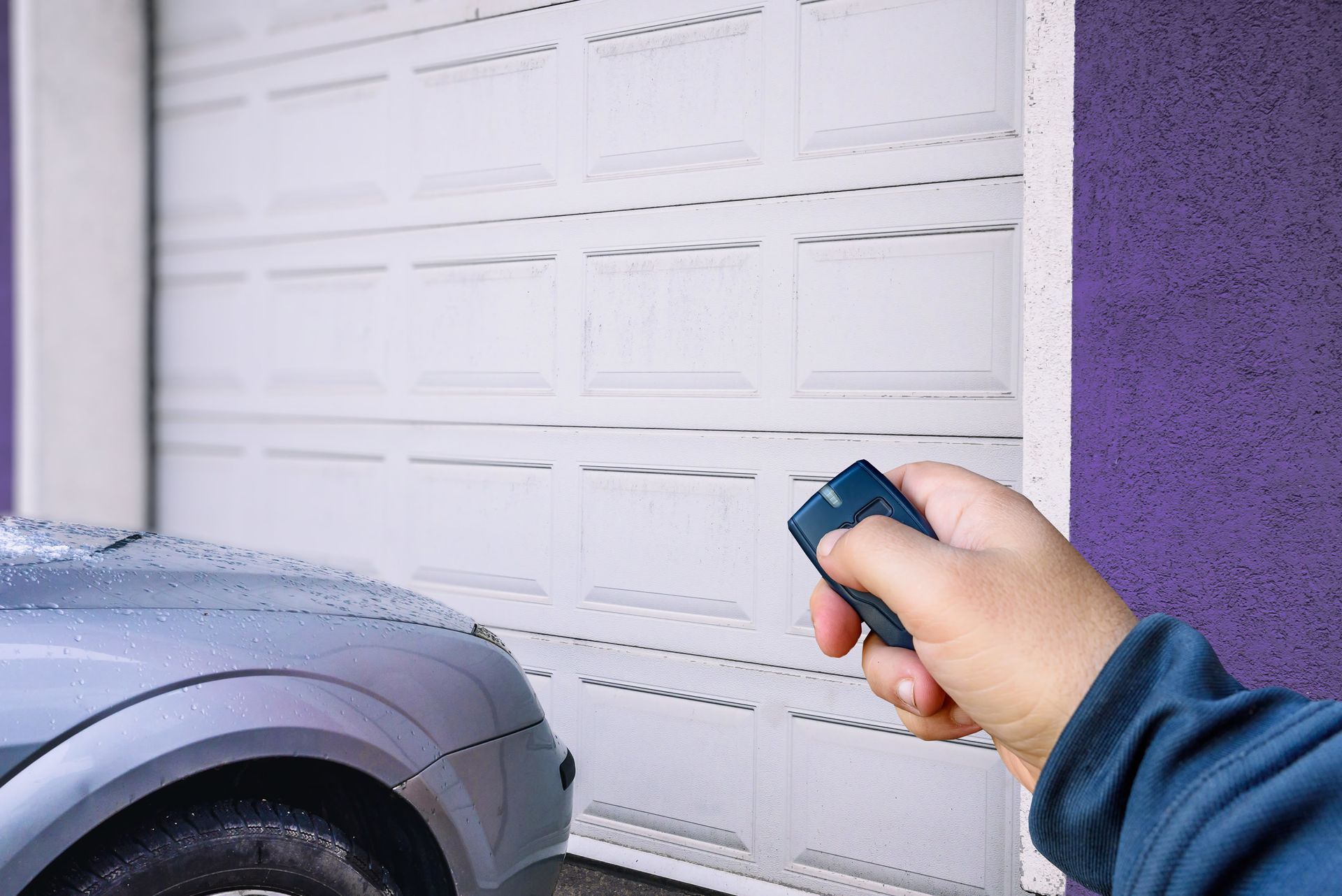 A hand holding a remote opens a white garage door next to a silver car.