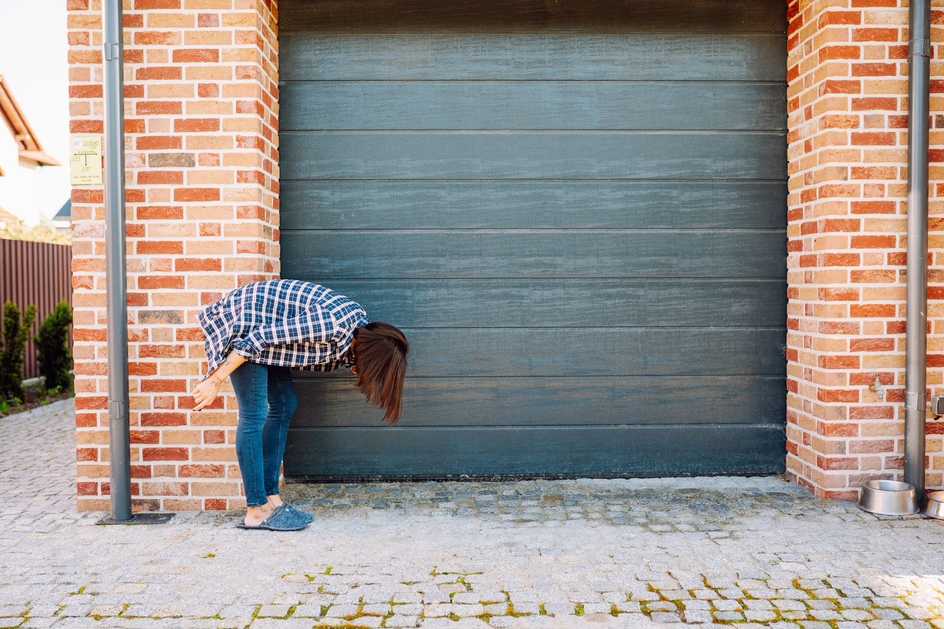 Person wearing plaid shirt and jeans looking down near garage door. Brick building.