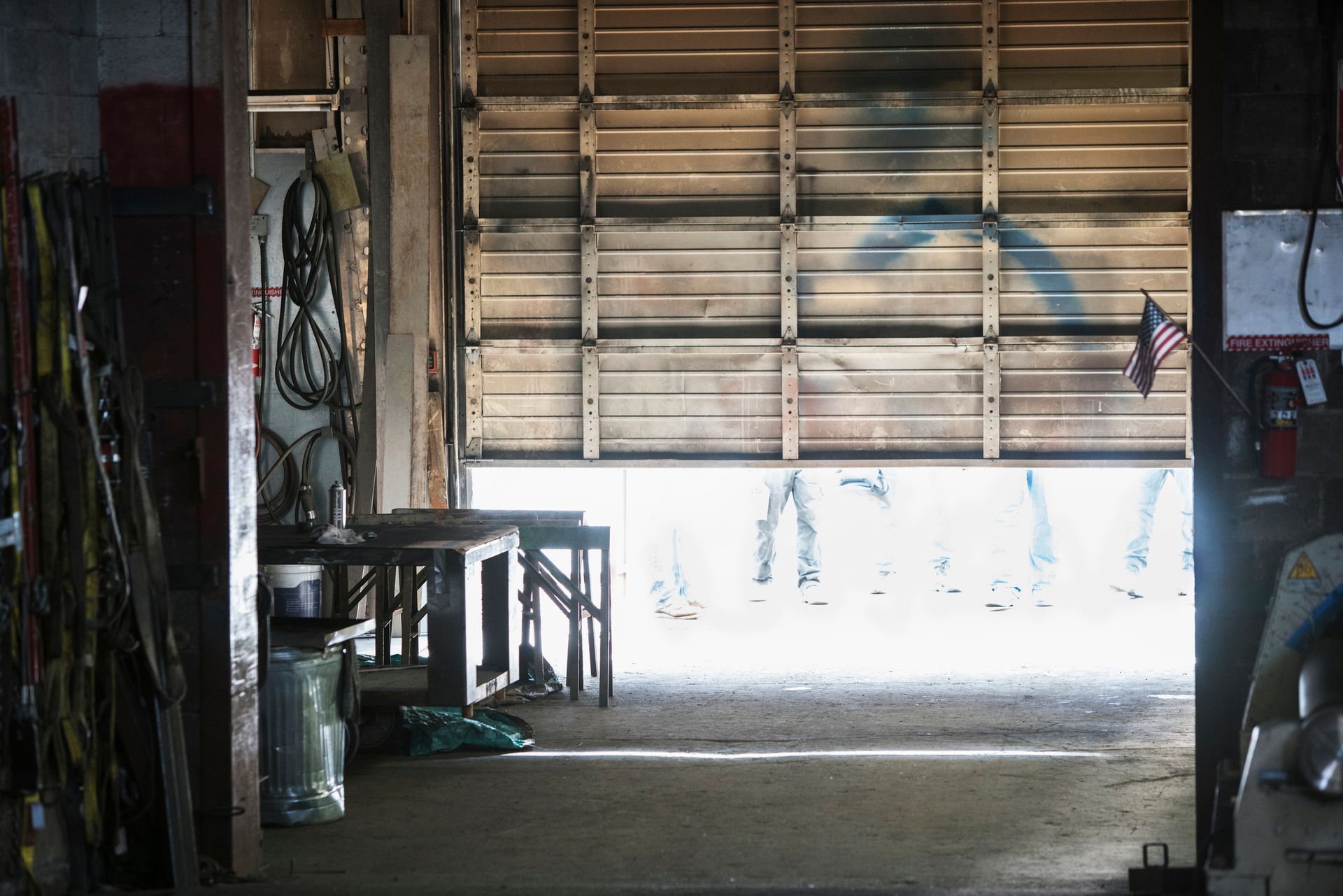 Inside a workshop, the partially open door reveals sunlight. Tables and equipment sit inside, an American flag hangs.