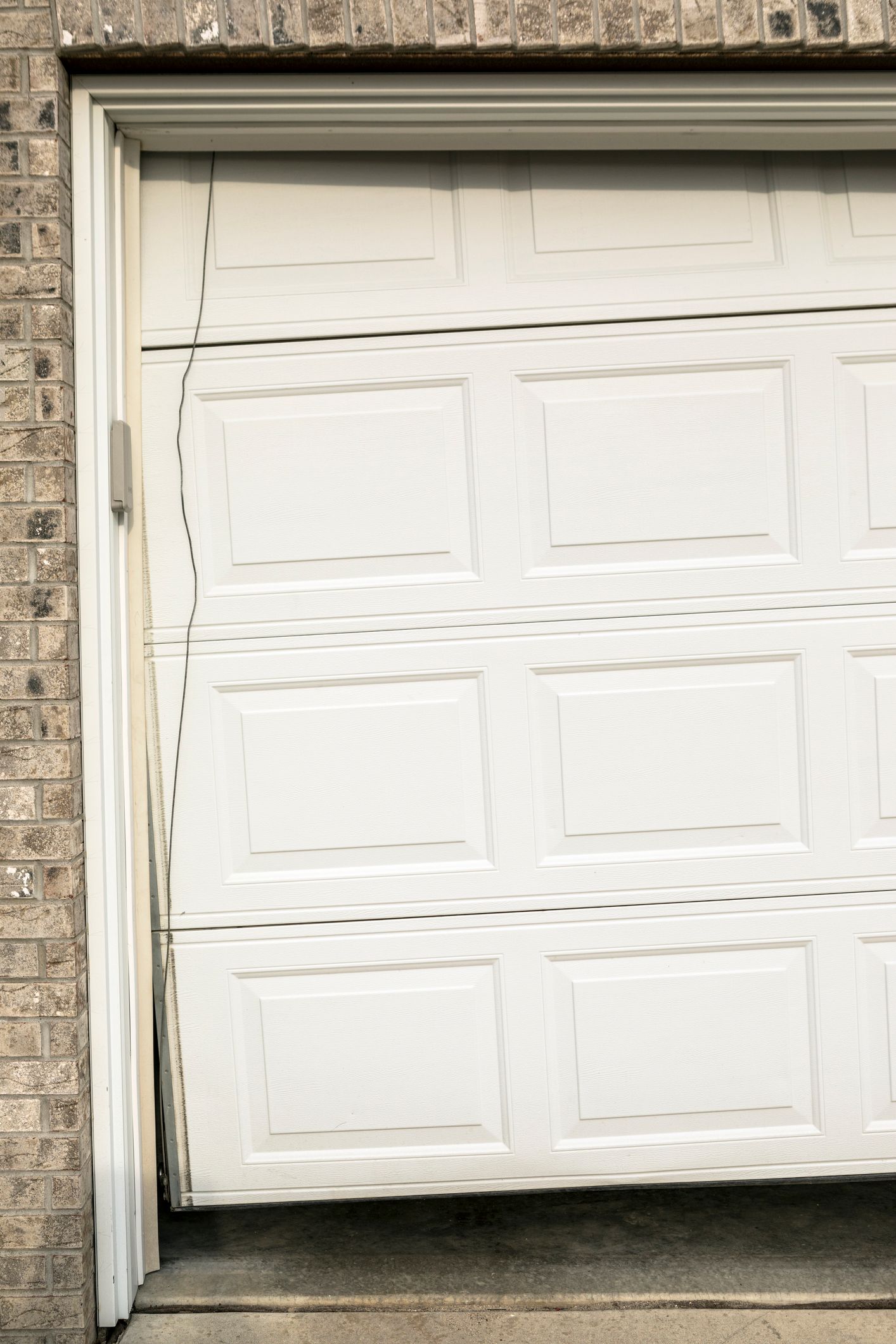 White garage door, partially open and misaligned in its frame, set against brick wall.