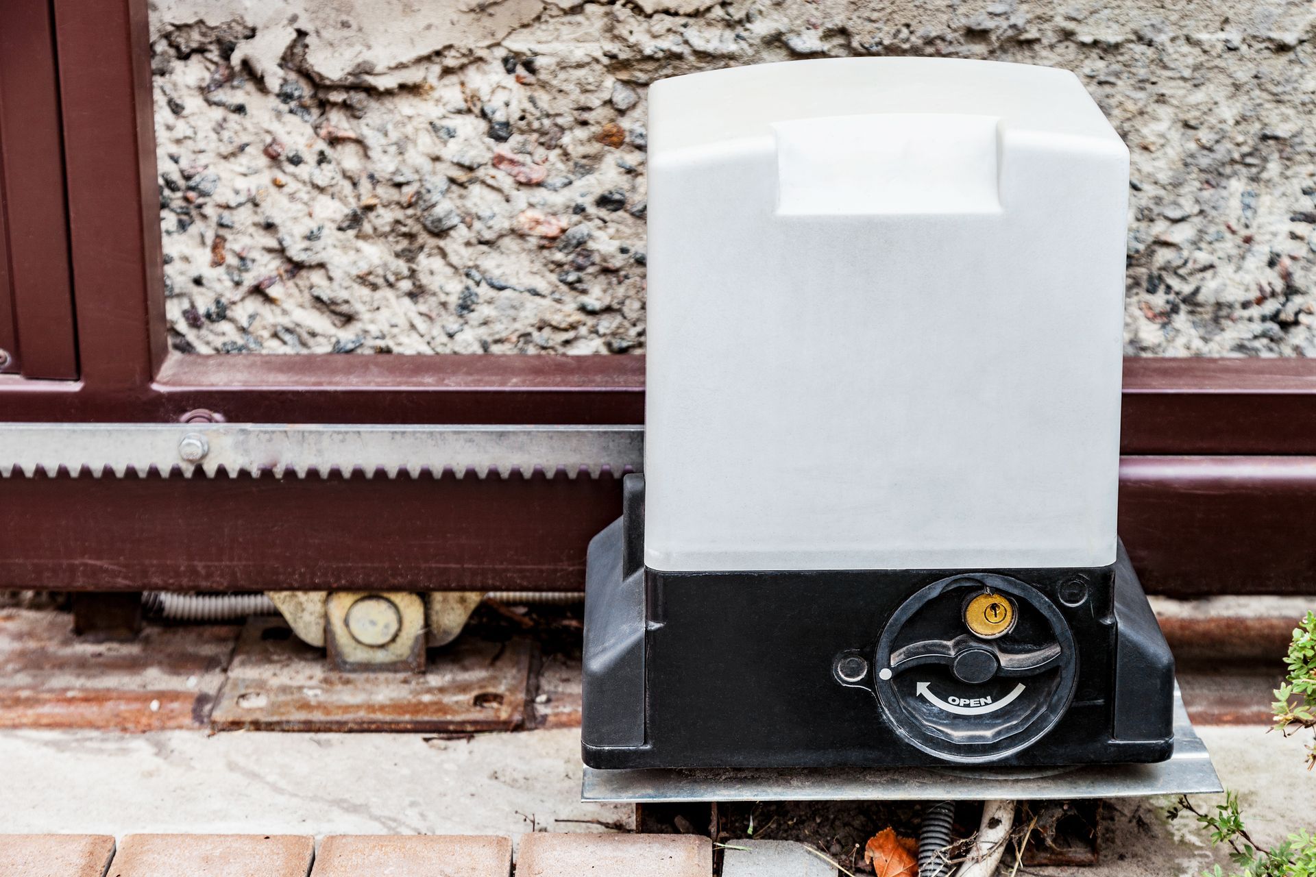 Sliding gate opener, black and white box, next to a brick wall and metal track.