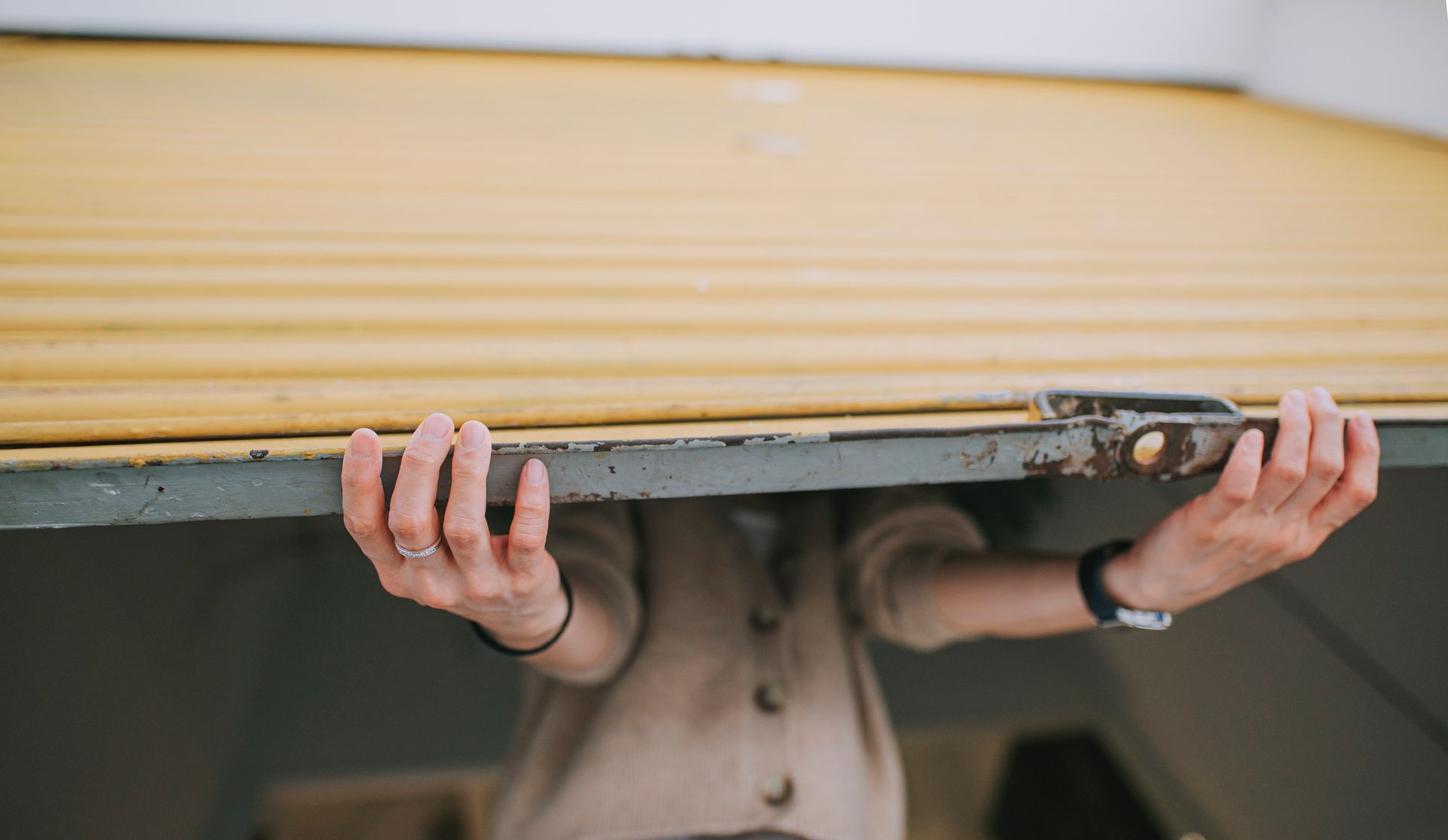 Person lifting a yellow roll-up garage door; hands visible, wearing a watch and ring.