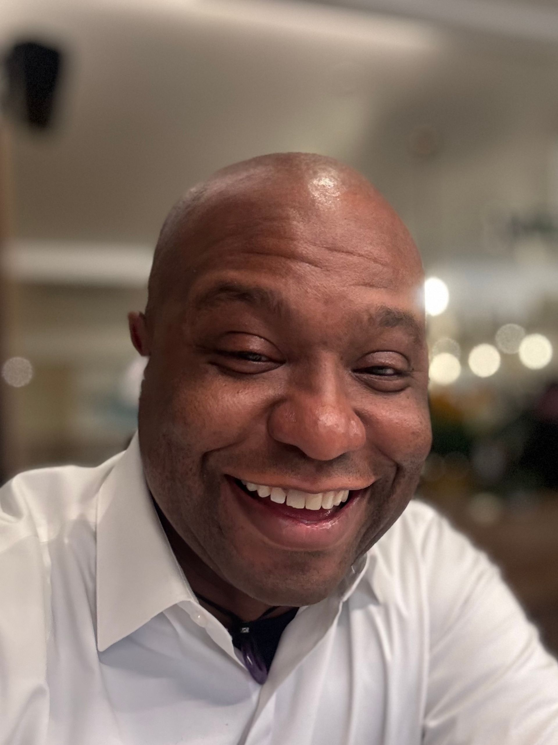 Smiling person in a white collared shirt, indoors, with blurred background lights.