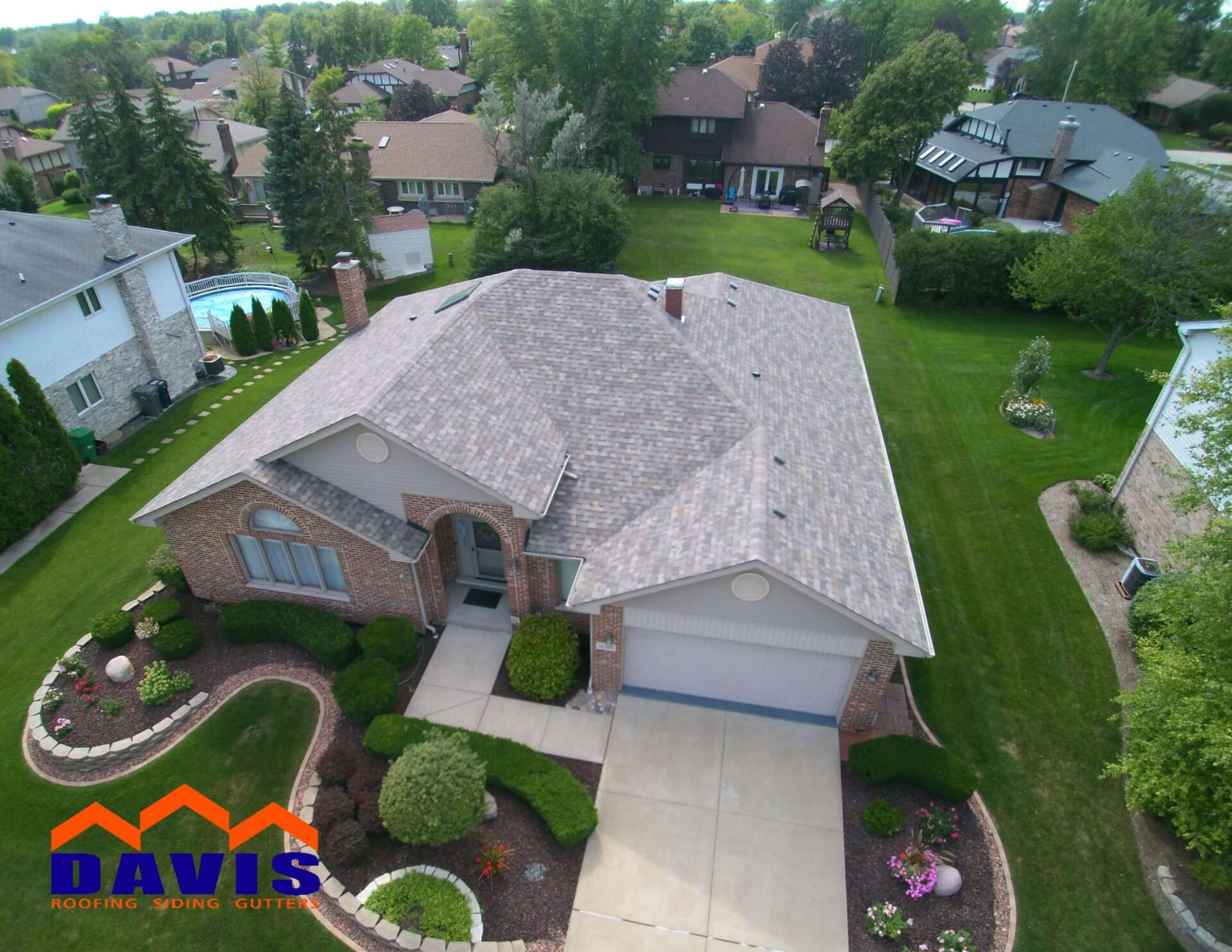 Aerial view of a brick home with a newly shingled roof, surrounded by green lawn and landscaping.