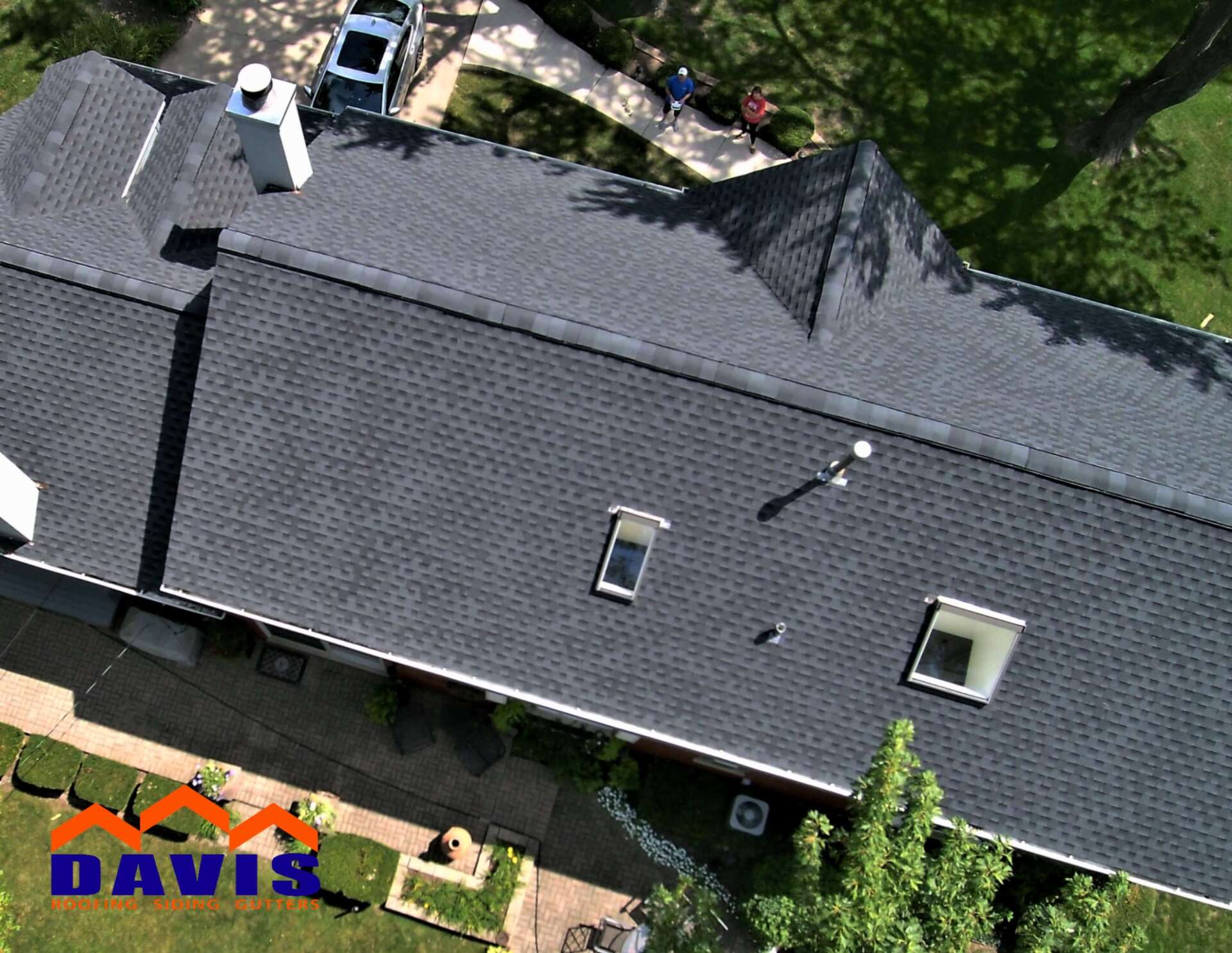 Aerial view of a dark gray shingled roof with skylights, a chimney, and workers; green lawn surrounds the house.
