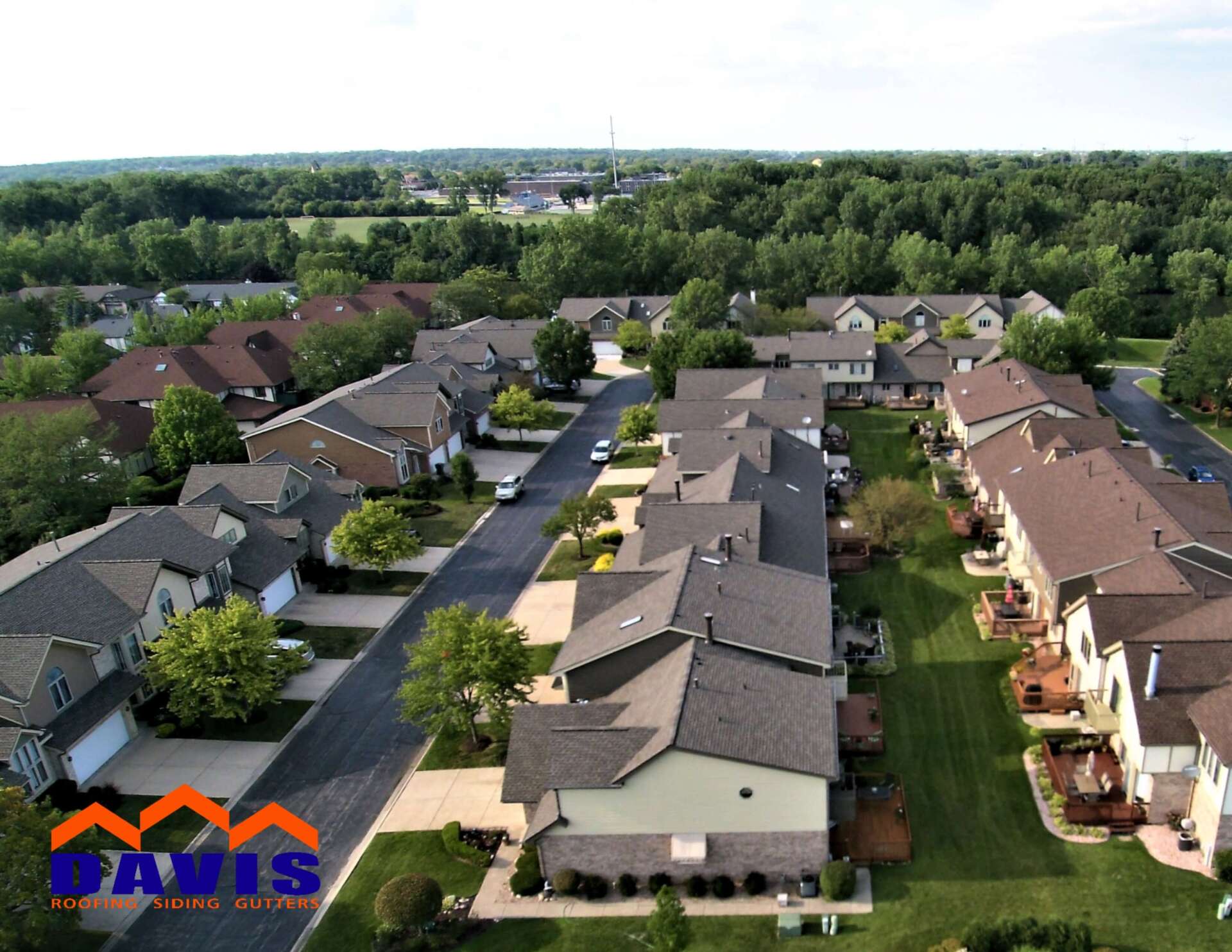 Aerial view of suburban townhomes with green lawns, trees, and a road on a sunny day.