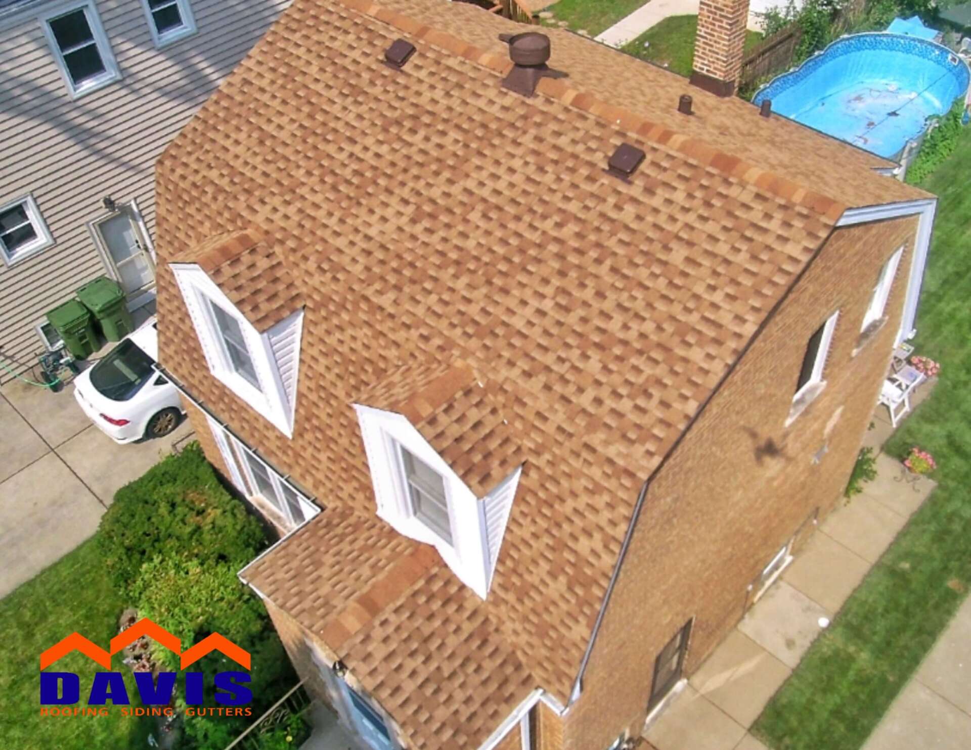 Brown shingled roof on a brick house with white trim, a car, and an above-ground pool in the background.