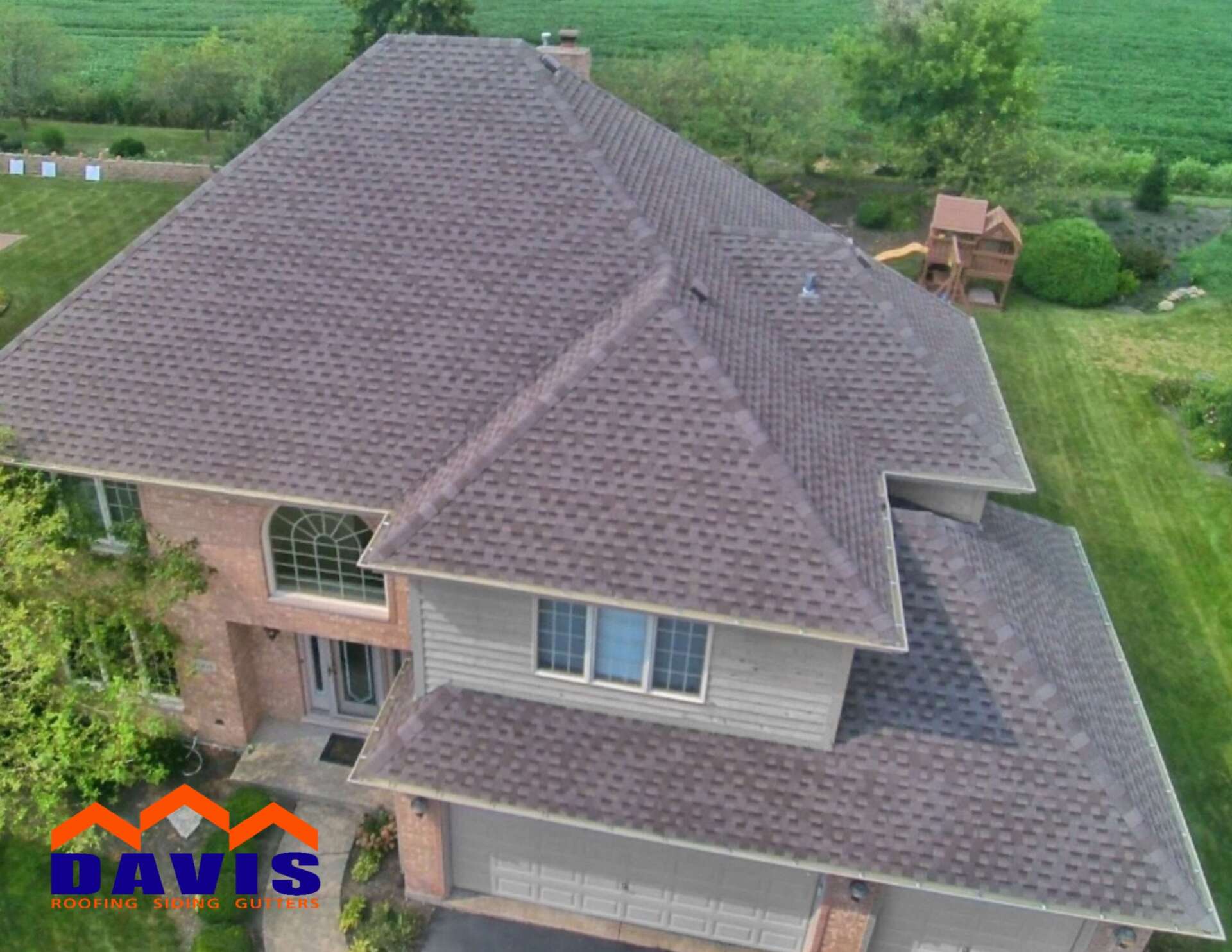Brown-shingled roof on a large house with a brick and siding exterior. Green grass surrounds it.