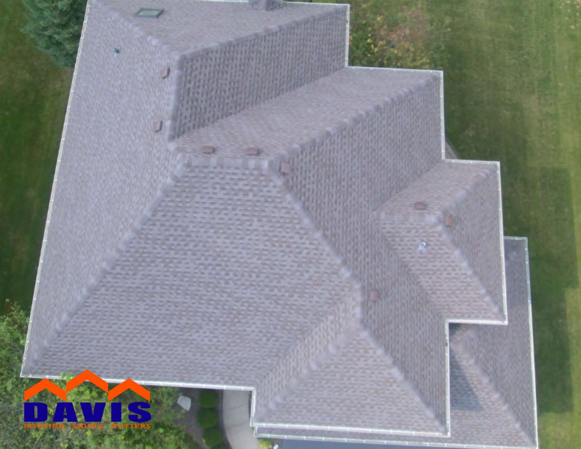 Overhead view of a gray shingled roof on a house, with a green lawn surrounding it. The logo of Davis Roofing is on the bottom left.