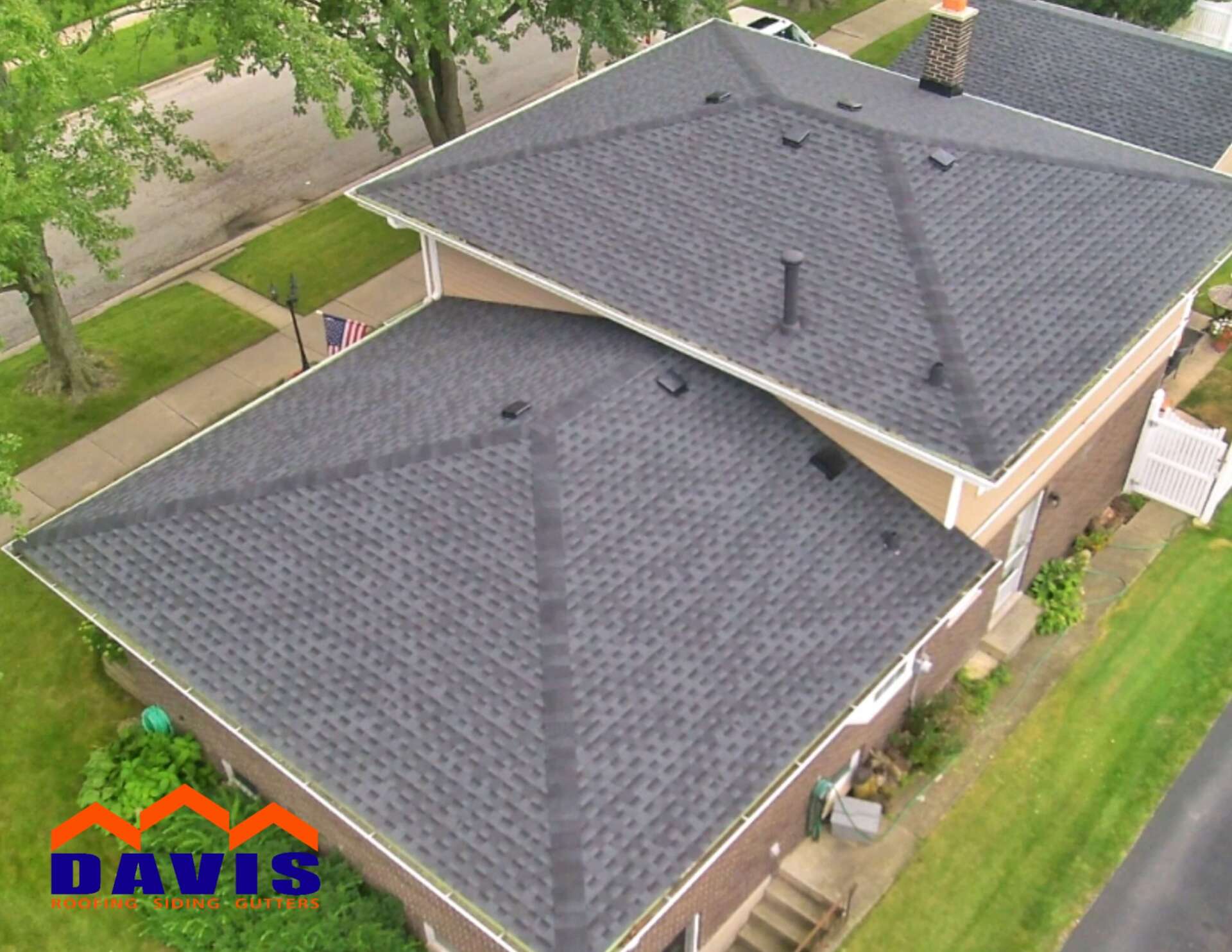 Aerial view of a house with dark gray shingled roofs. The house is surrounded by green lawn and trees.
