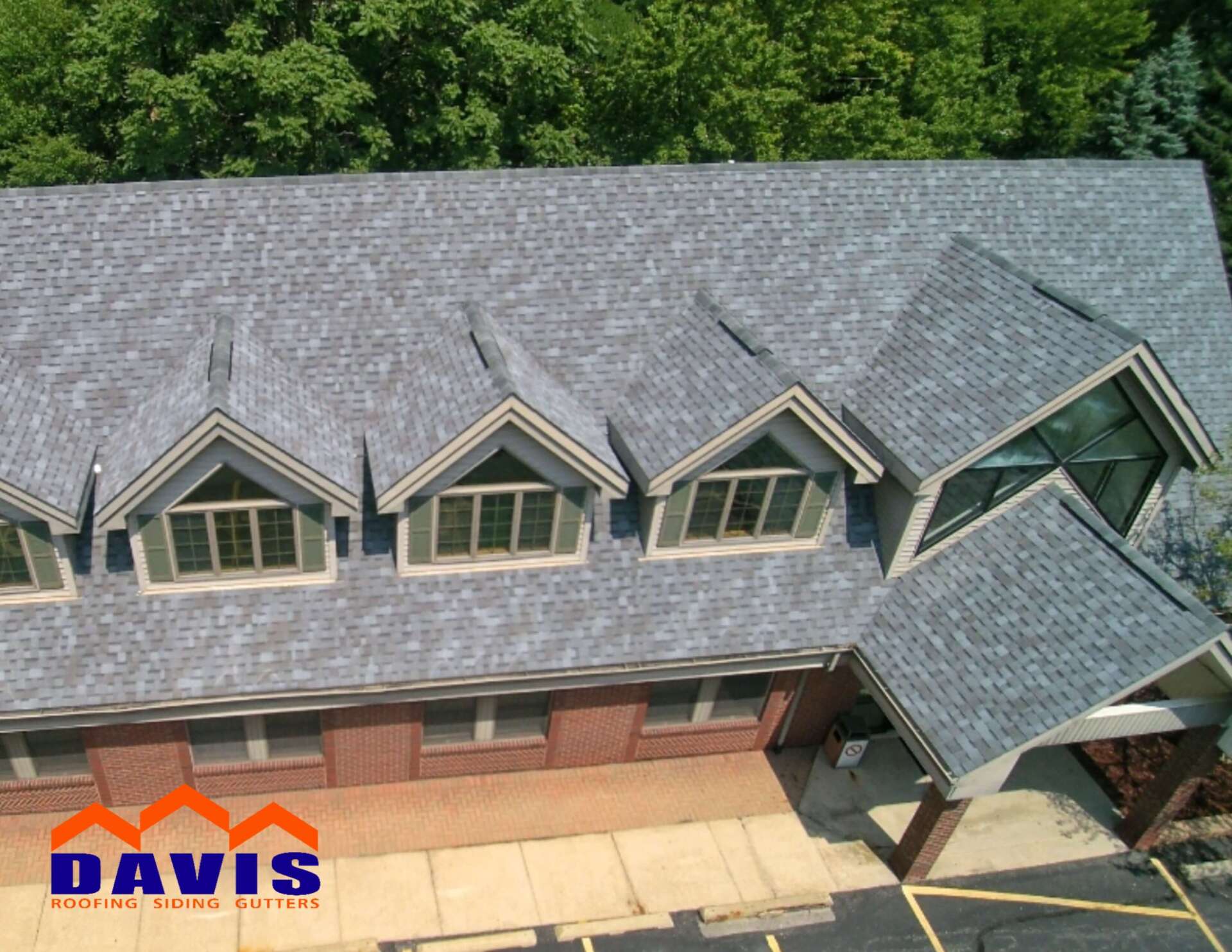 Aerial view of a building with a gray shingle roof and multiple dormers. Brick facade, surrounded by trees.