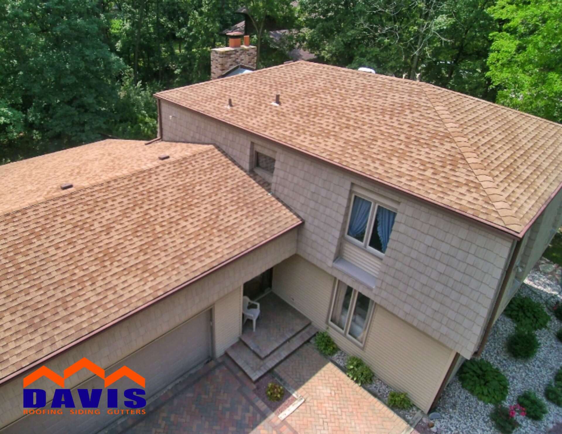 Aerial view of a house with a new brown shingle roof, beige siding, and surrounding green trees.