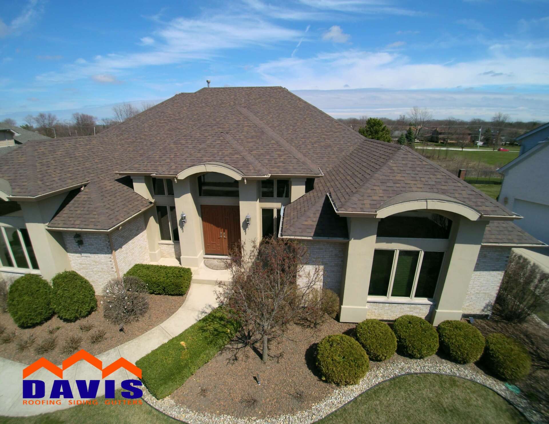 Aerial view of a house with brown roof and tan exterior; green lawn; blue sky.