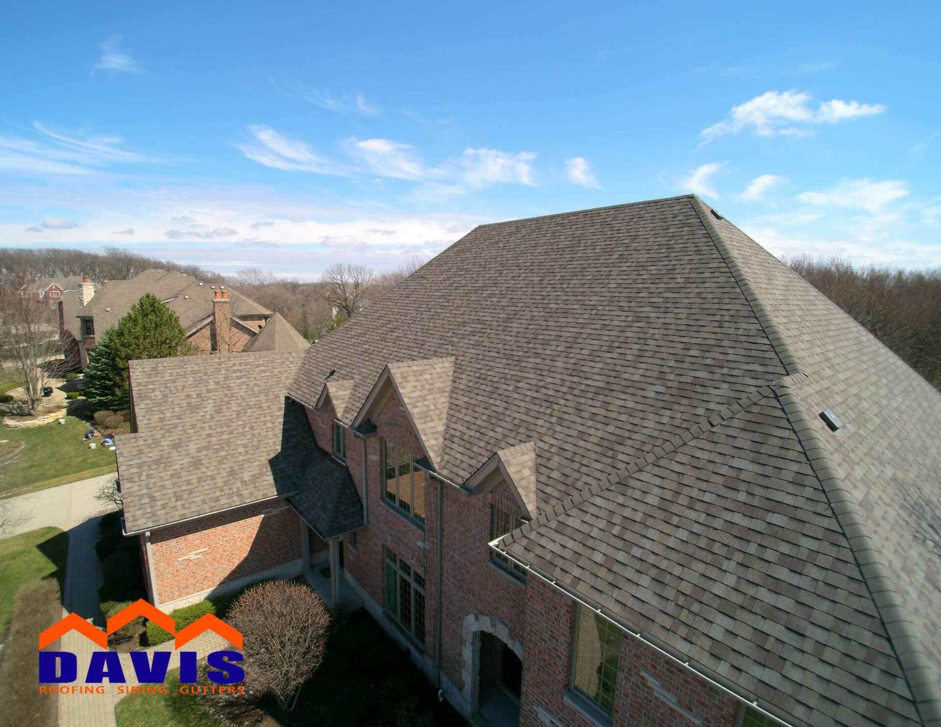 Aerial view of a brick house with a dark gray shingle roof, trees, and blue sky. 