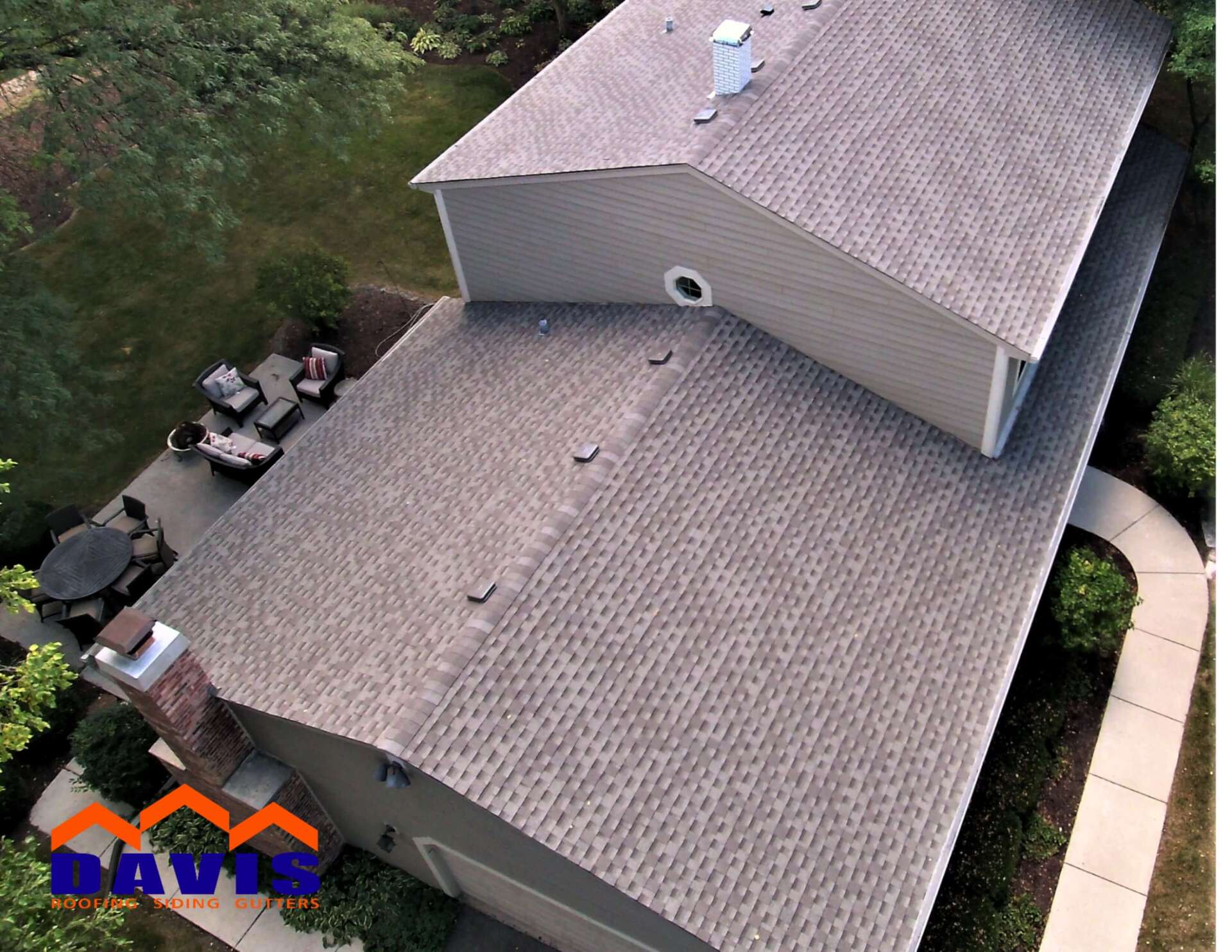 Overhead view of a house with brown shingle roof. Attached tan structure and patio with seating are visible.