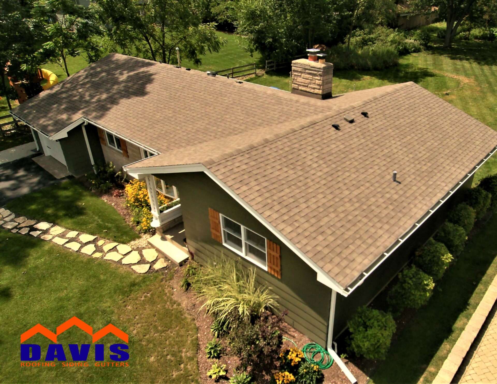 Brown-roofed house with green siding, chimney, and well-maintained landscaping on a sunny day.
