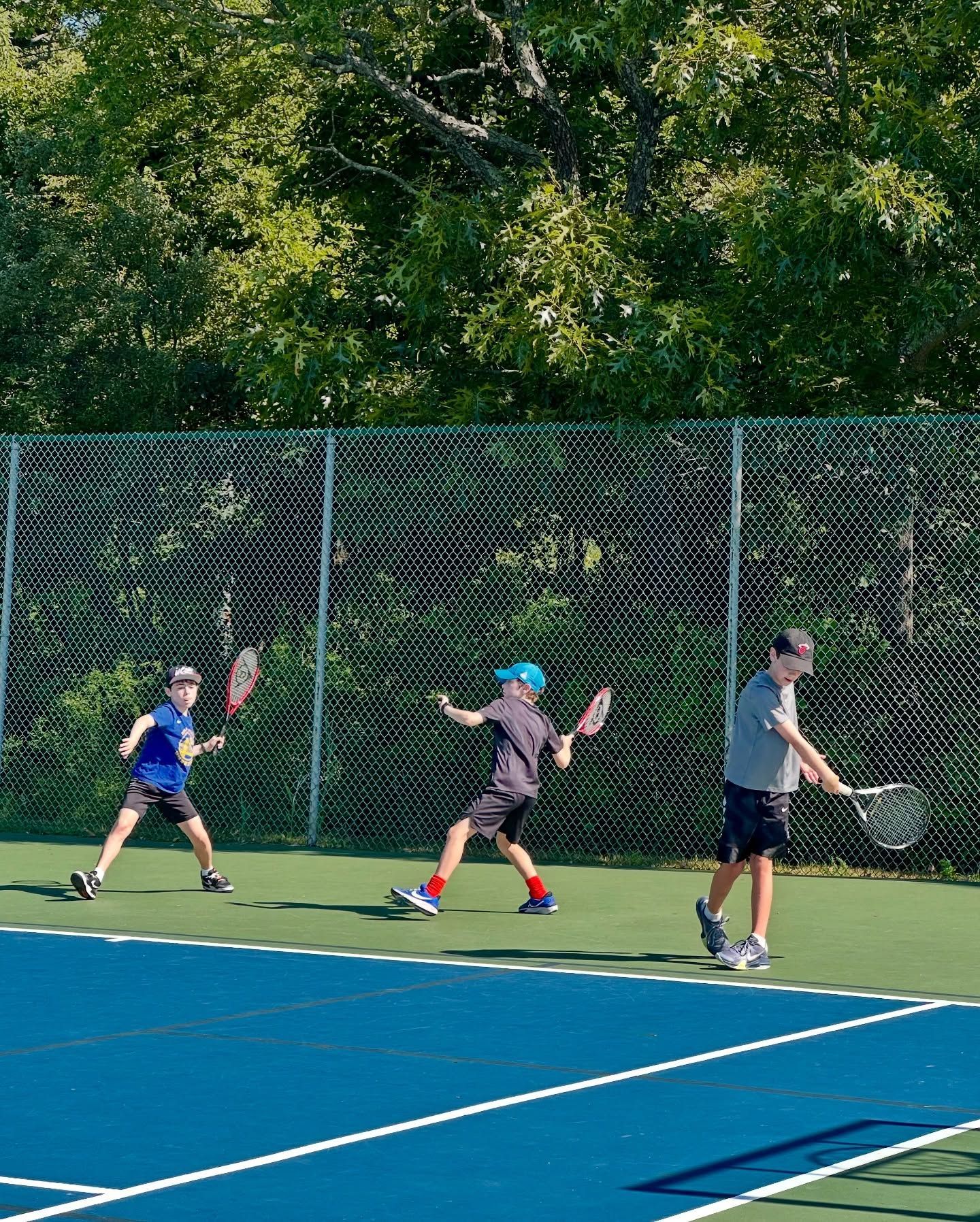 Three kids playing tennis on a blue court. One swings, one reaches, and one prepares. Green trees in the background.