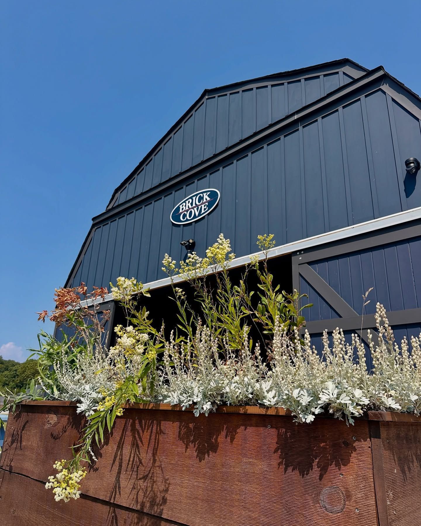 Dark blue barn-like building with flowers in a planter.