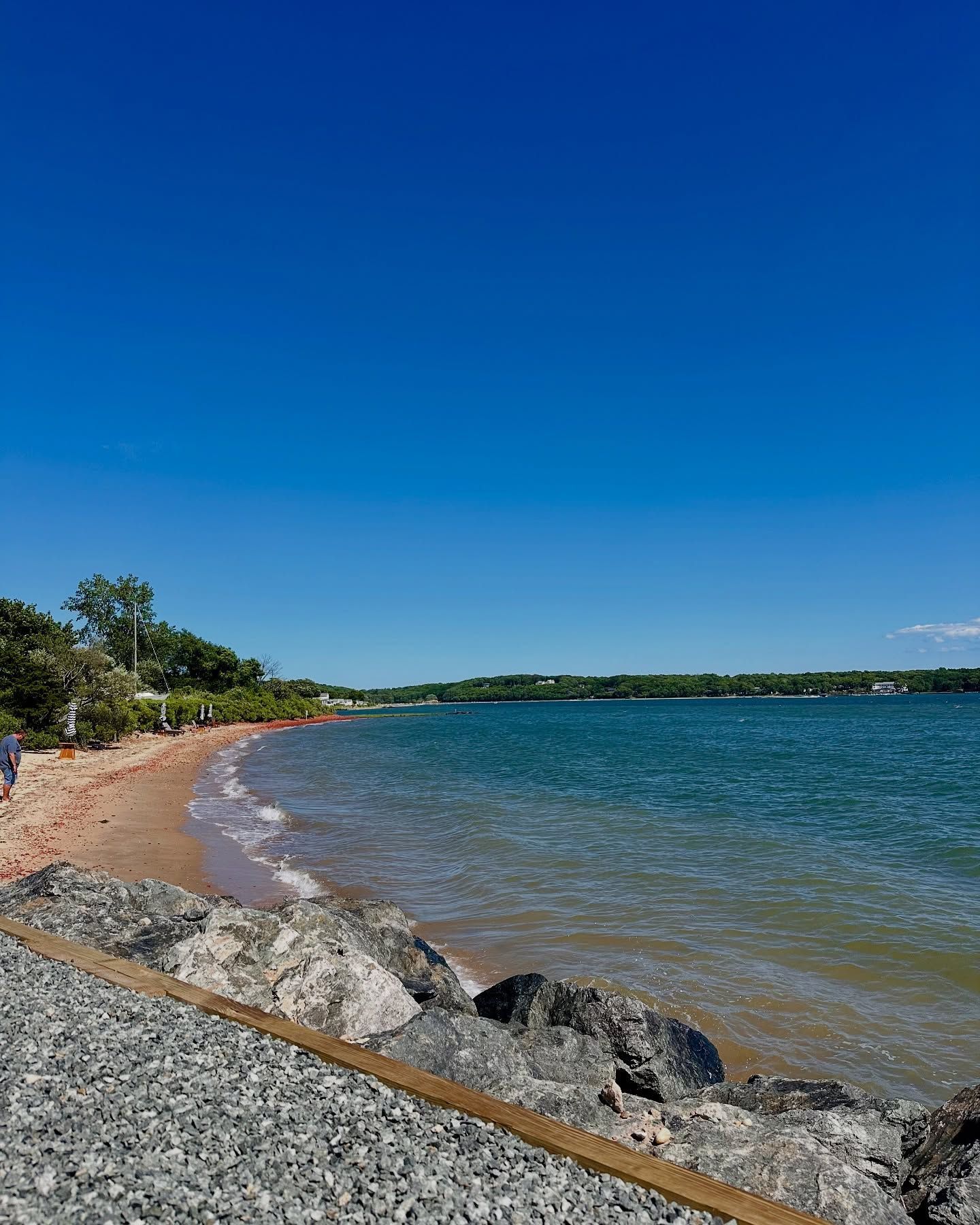 Bright blue sky over a sandy beach and calm, blue water. Large rocks in the foreground, trees on the horizon.