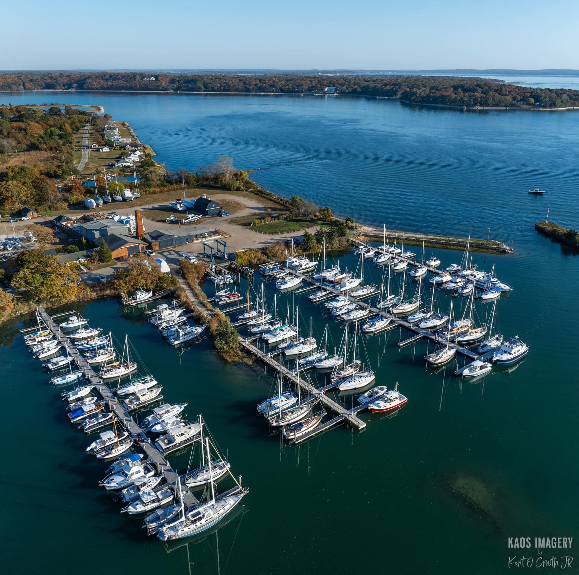Aerial view of a marina with sailboats docked; blue water, fall foliage, buildings on the shore.