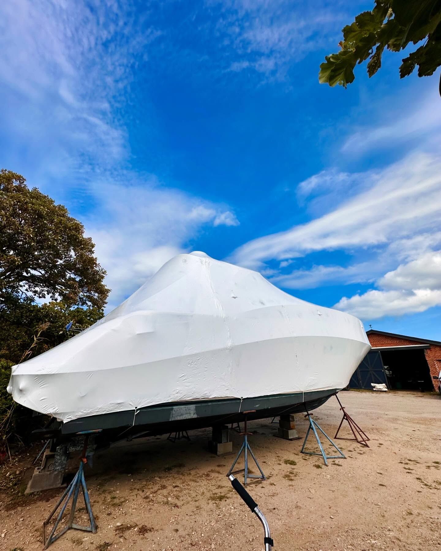 A boat covered in white tarp on stands outside under a bright blue sky.