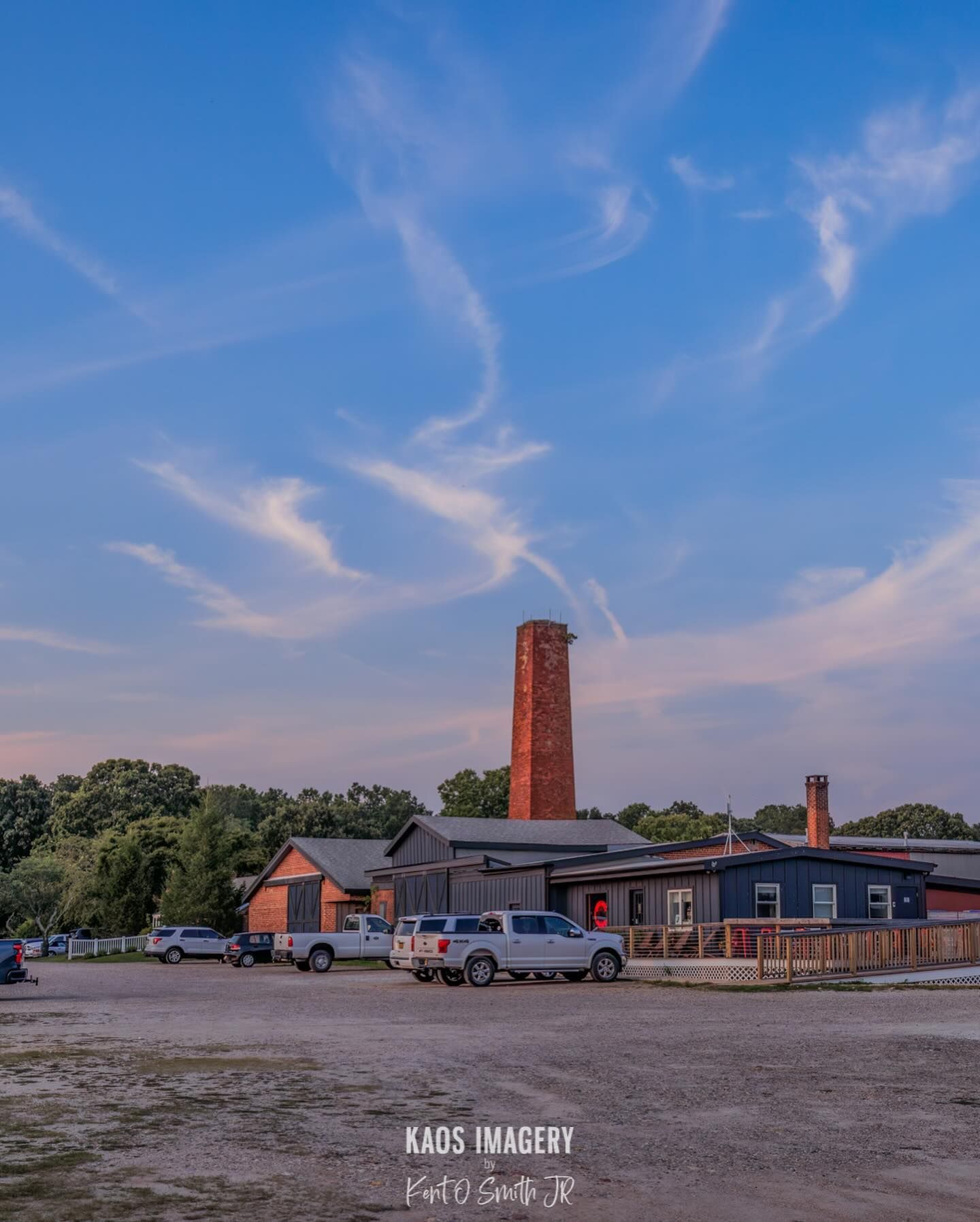A brick smokestack rises above a factory. Cars parked outside with a blue and pink sky.