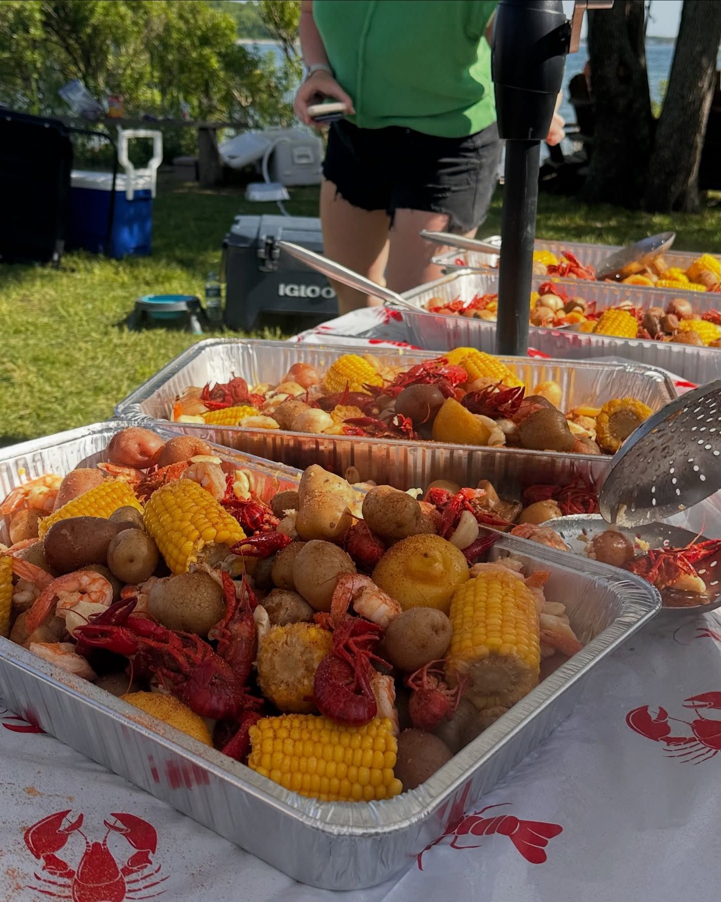 Crawfish boil with corn, potatoes, and shrimp in metal trays on a table with a crawfish-themed tablecloth; outdoor setting.