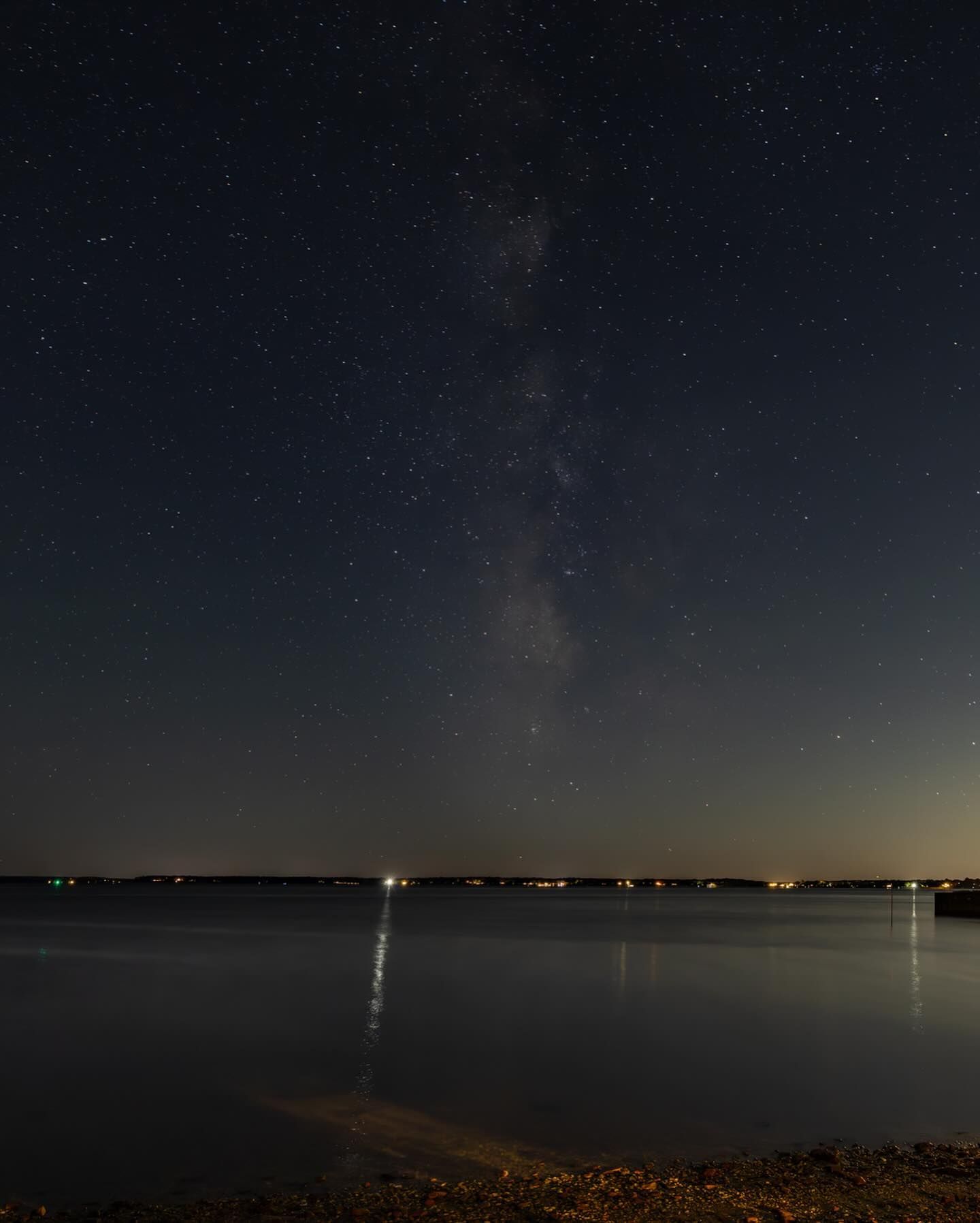 Milky Way over a calm lake, with lights on the distant shore under a dark blue sky.