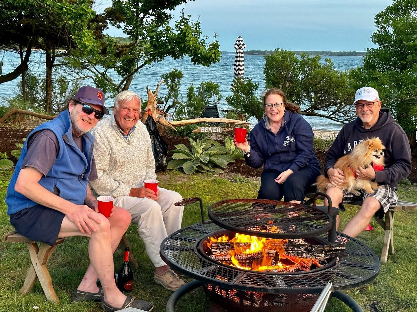 Four people gathered around a fire pit near water, smiling, holding drinks, one with a small dog.