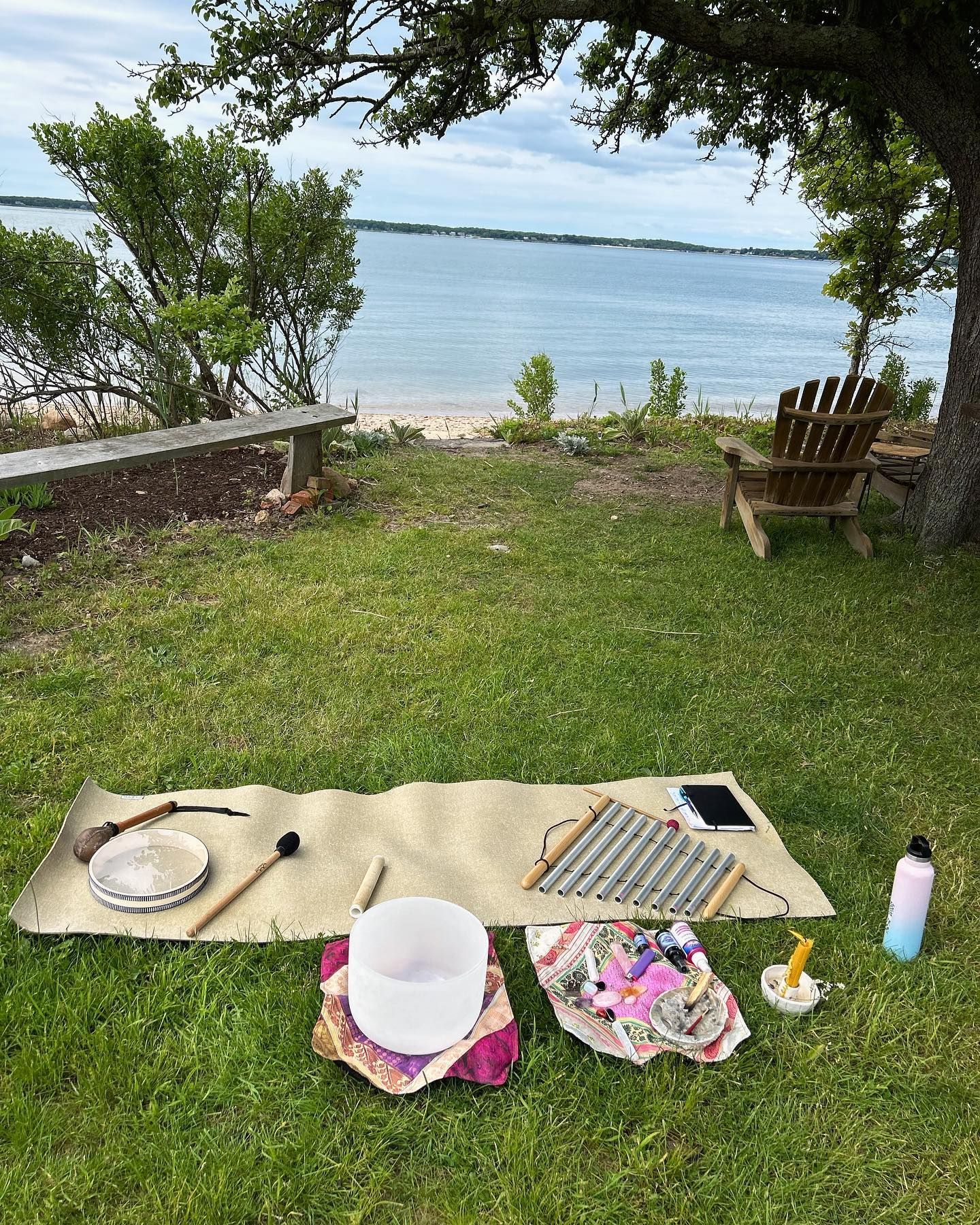 Outdoor sound healing setup with instruments on a mat overlooking the water and a bench.
