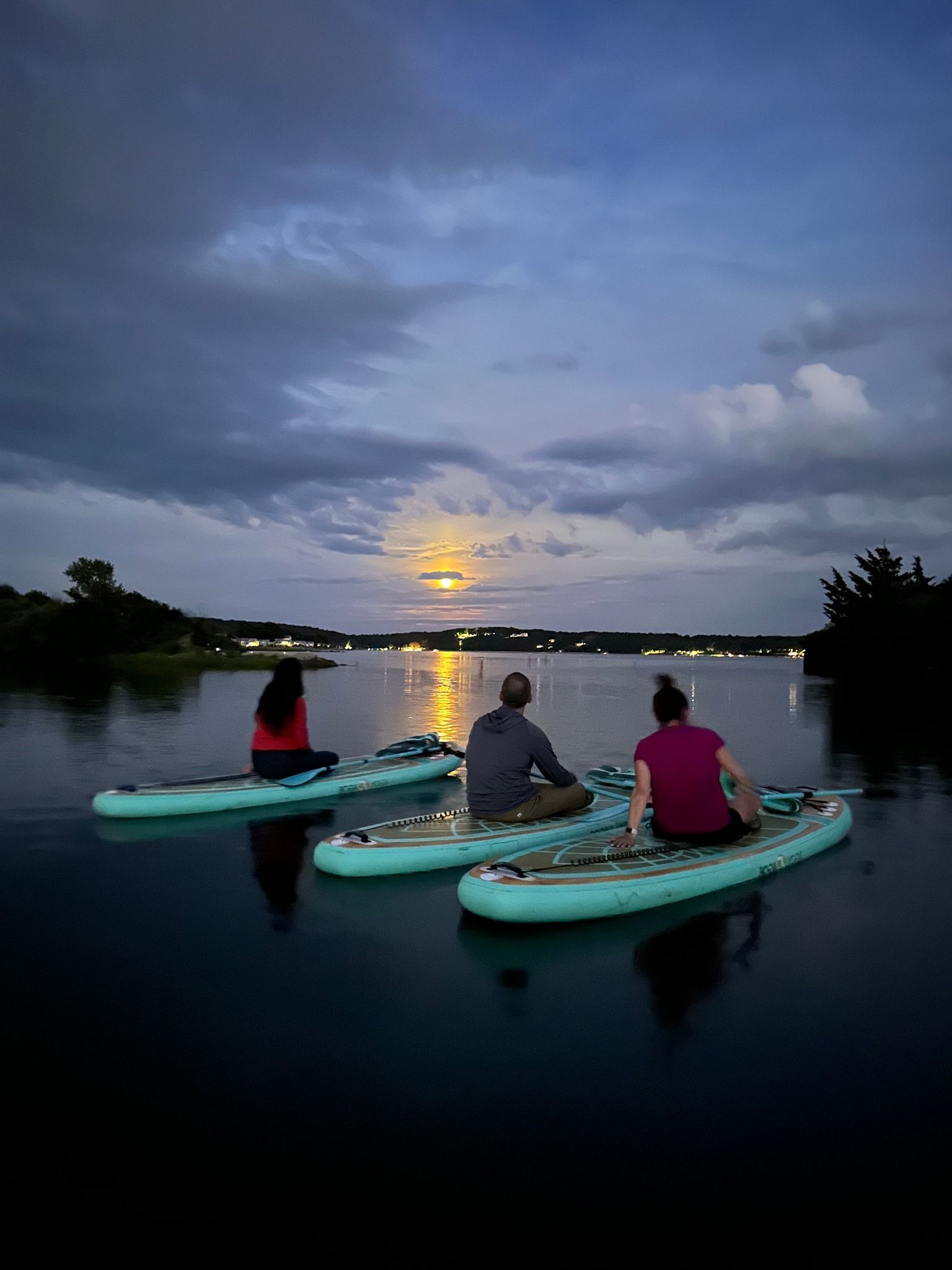 Three people on paddleboards at dusk, watching the moon rise over the water.