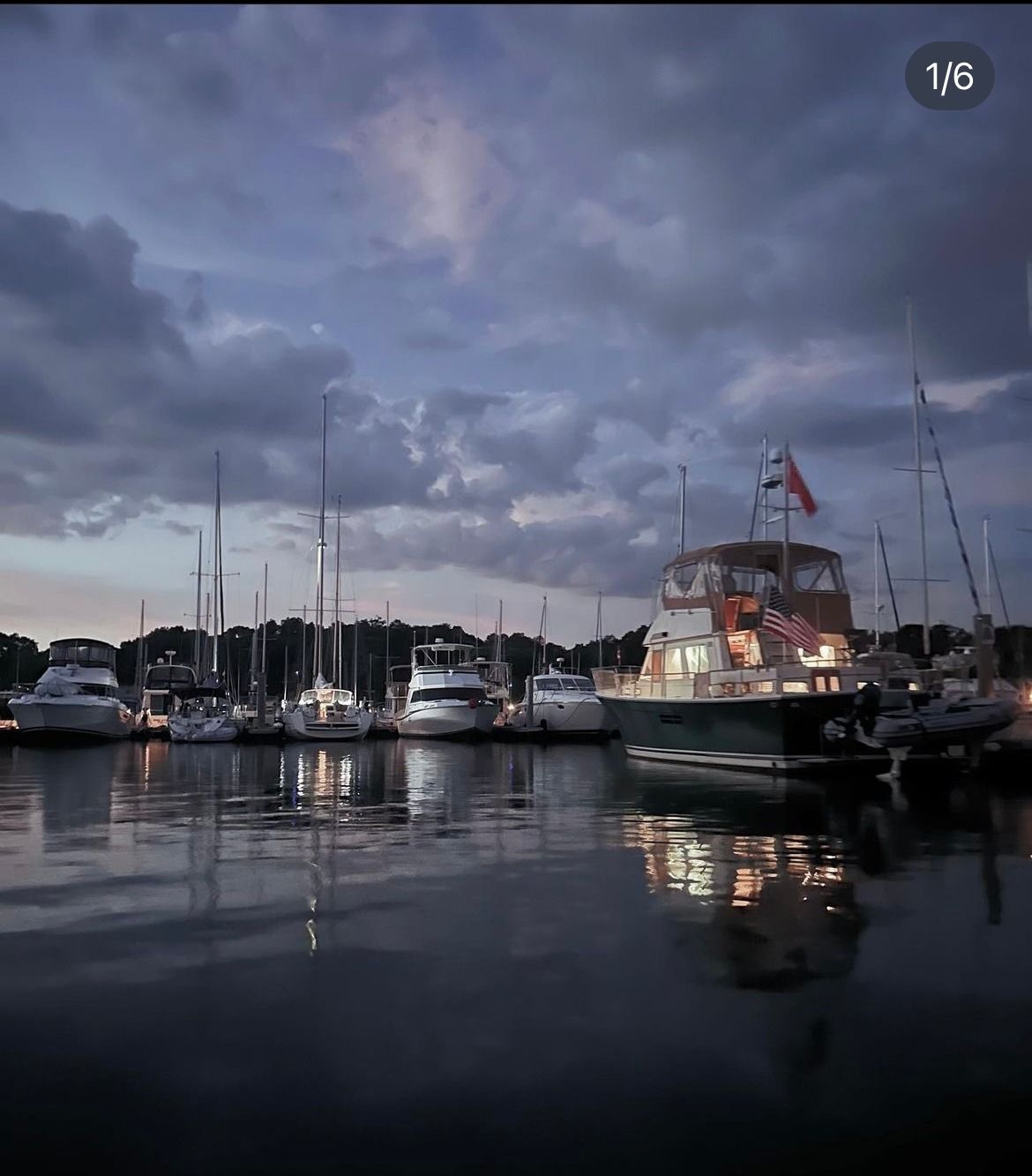 Boats docked in a harbor at dusk, with dark water reflecting the lights and cloudy sky.
