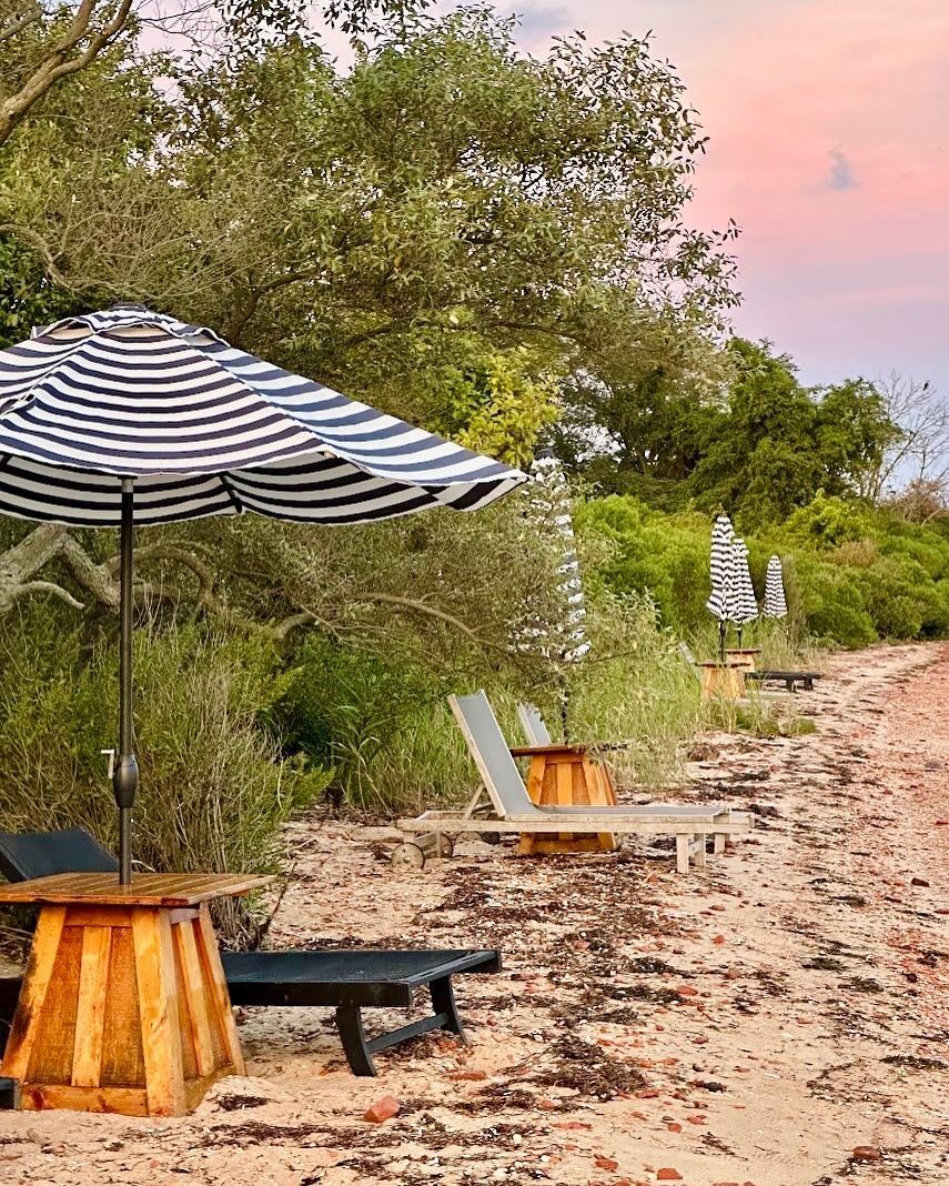 Beach scene with striped umbrellas, lounge chairs on a sandy shore, and green foliage in the background.