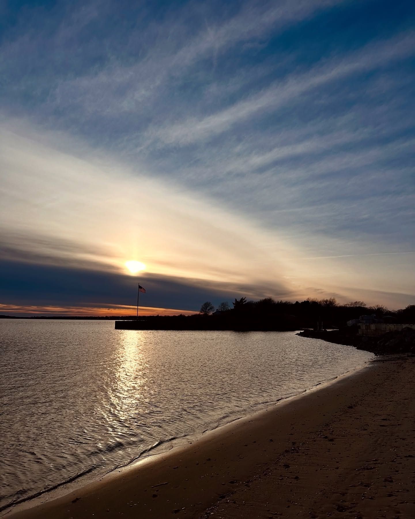 Sunset over water, silhouetted shoreline, dock, and cloud-streaked sky.