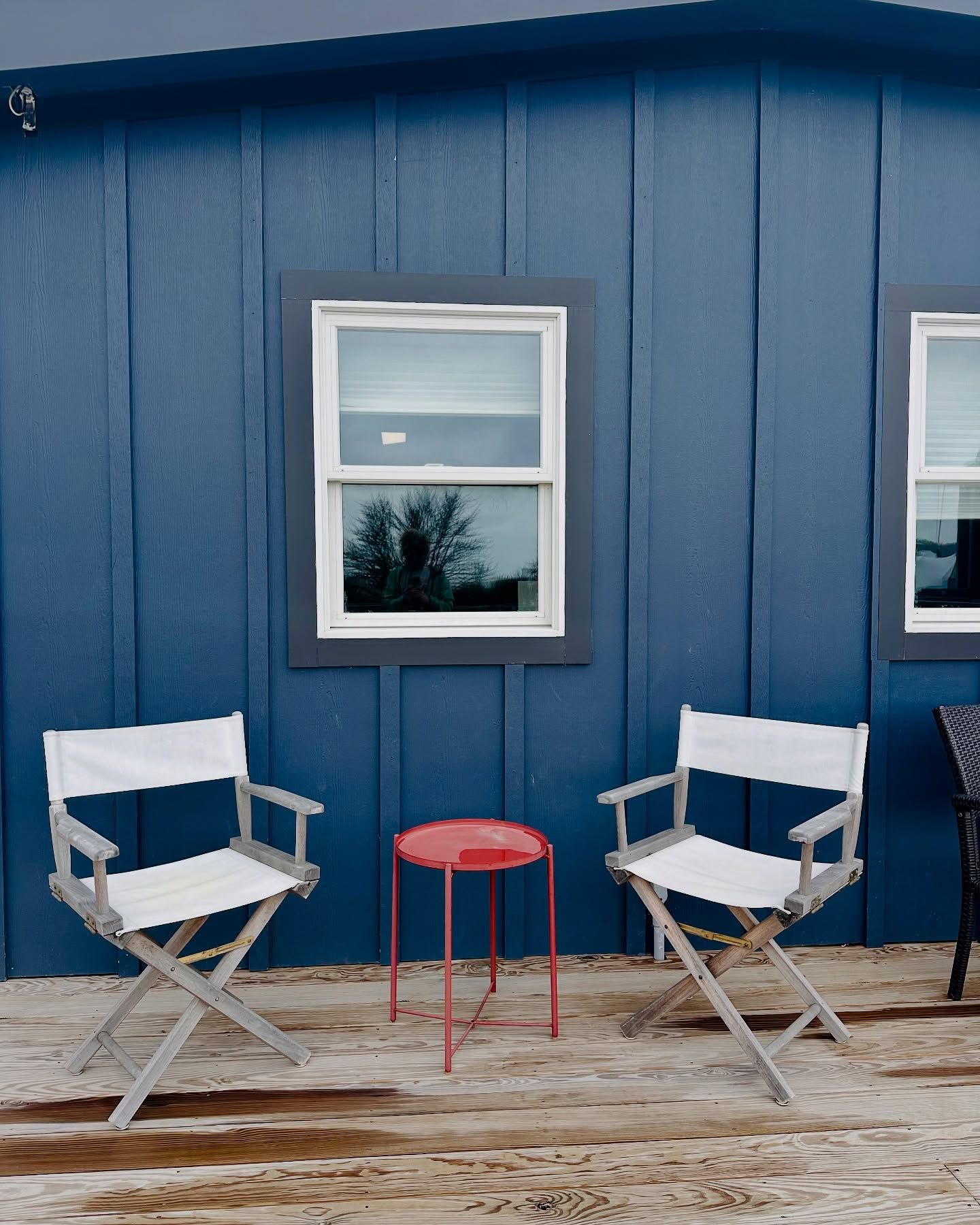 Two white director's chairs and a red stool on a porch, in front of a blue building with windows.