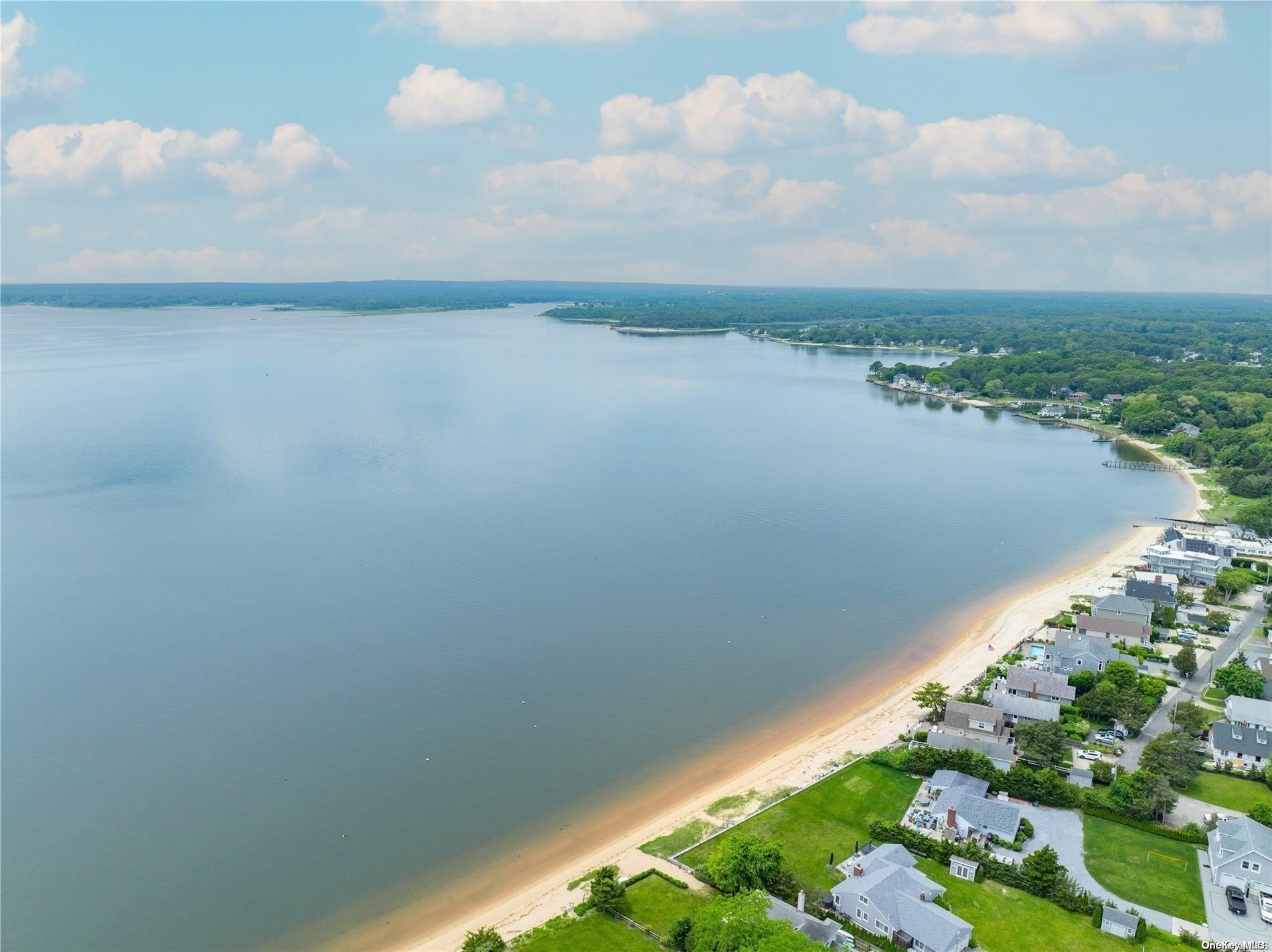 Aerial view of a sandy beach, houses, and a calm body of water under a blue sky with clouds.