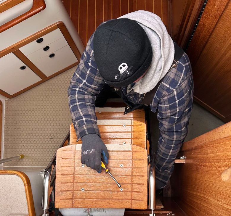 Person working on a wooden boat step, using a screwdriver. They wear gloves, a hat, and plaid shirt.