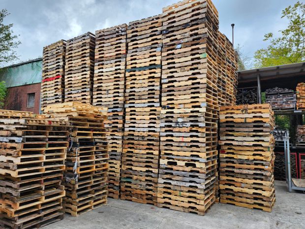 Stacks of wooden pallets outdoors, varying sizes and heights, mostly brown, some with colored markings.