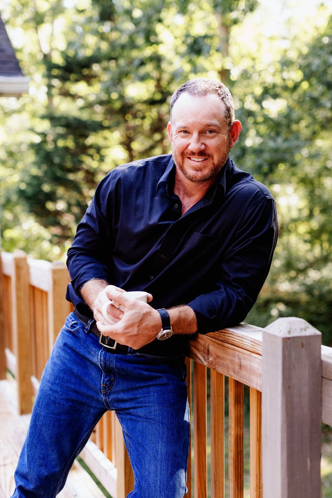 Man in black shirt and jeans leans on wooden deck railing, smiling.